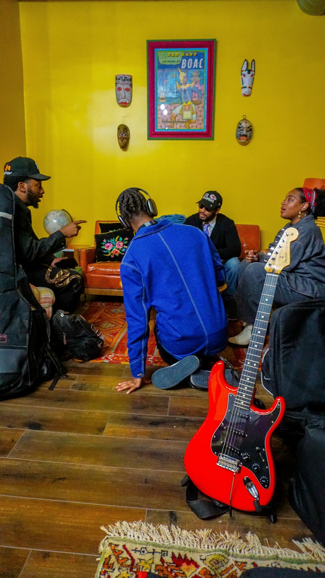 Group of people sitting and standing in a room with yellow walls, decorated with framed pictures and masks, wooden floor, red guitar in foreground.