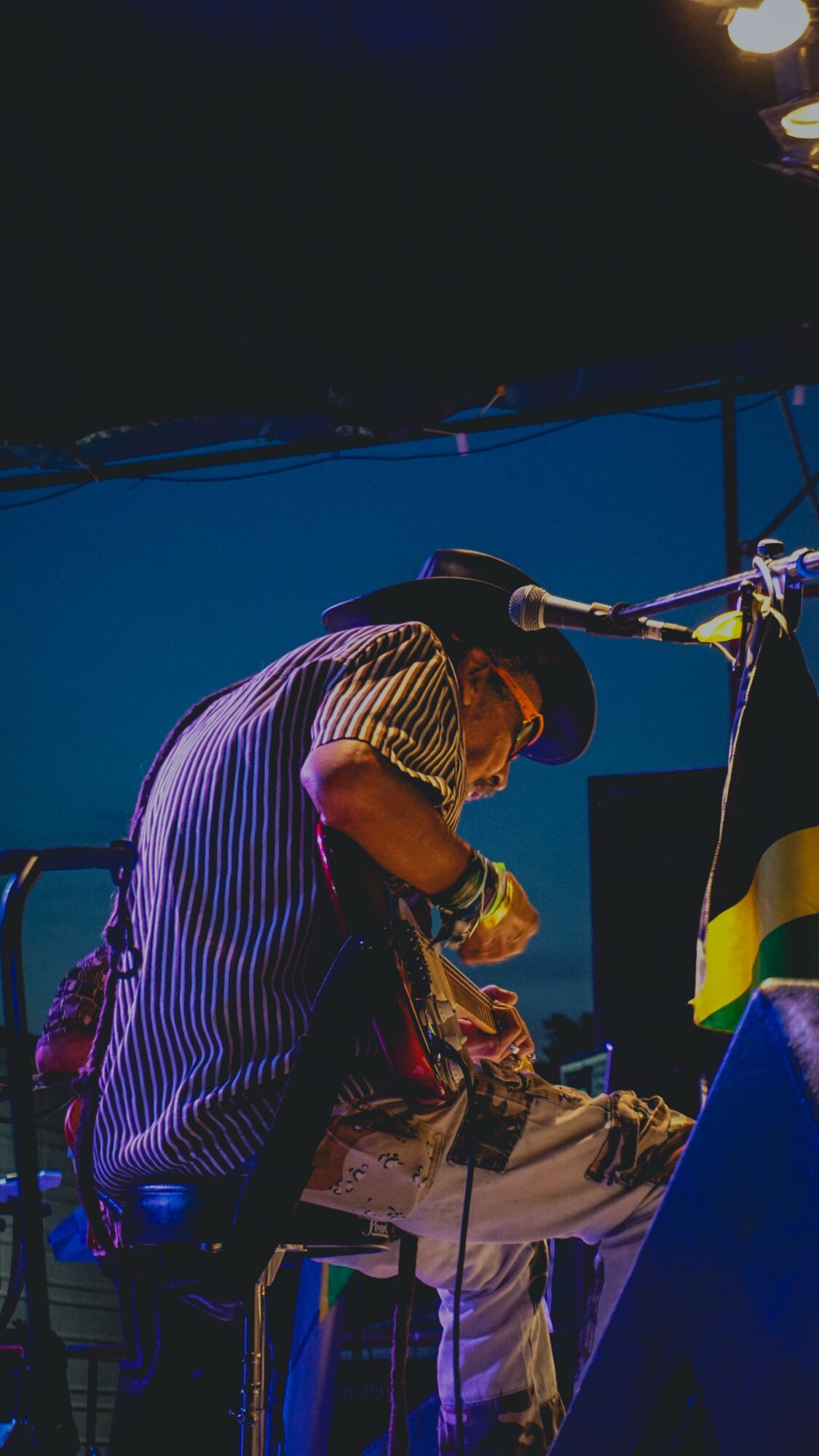 Person wearing a striped shirt and hat, sitting at a table with a microphone nearby, in a dimly lit setting.
