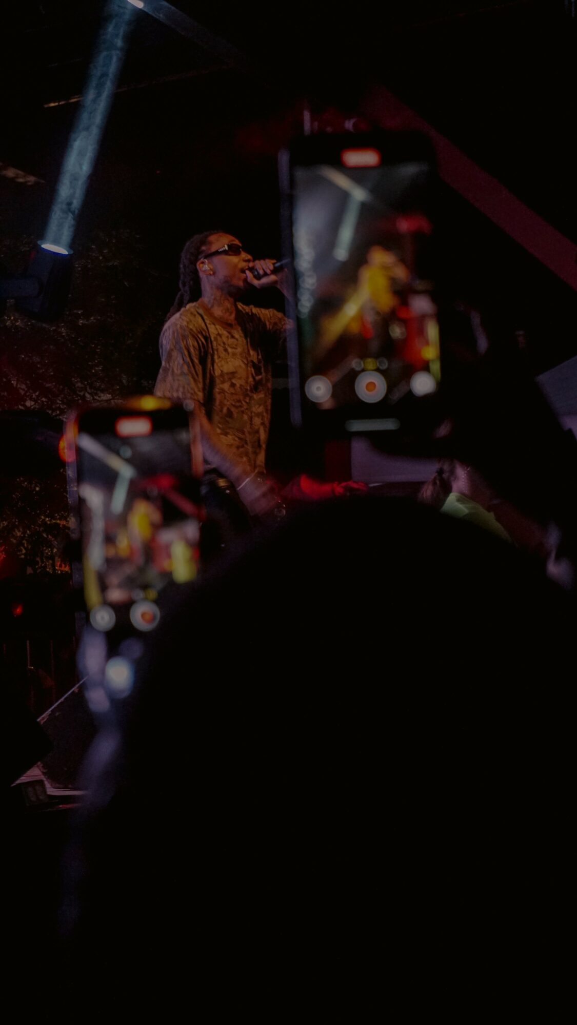 Performer on stage with microphone, audience recording with phones, dark background, colorful lights.