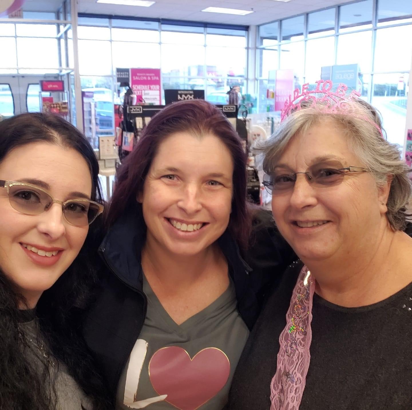 Three women smiling indoors with large windows and store displays in background.