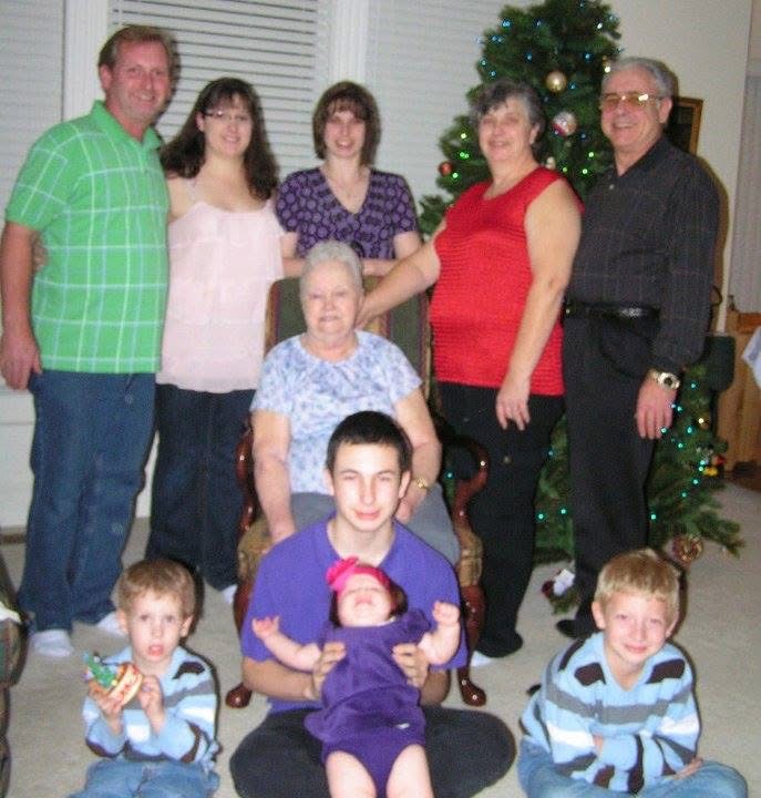 Group of seven people, including children, adults, and an elderly woman, gathered indoors near a decorated Christmas tree.