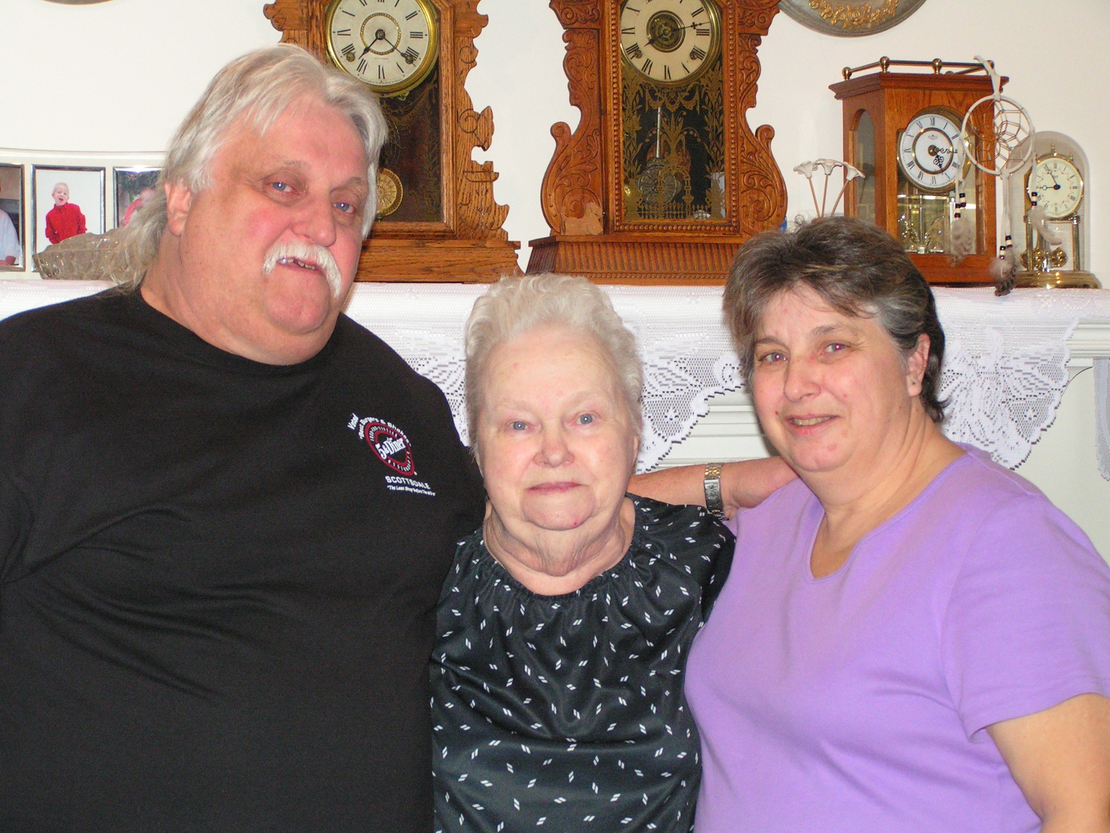Three people, two women and one man, standing indoors in front of a wooden cabinet with clocks and decorative items.