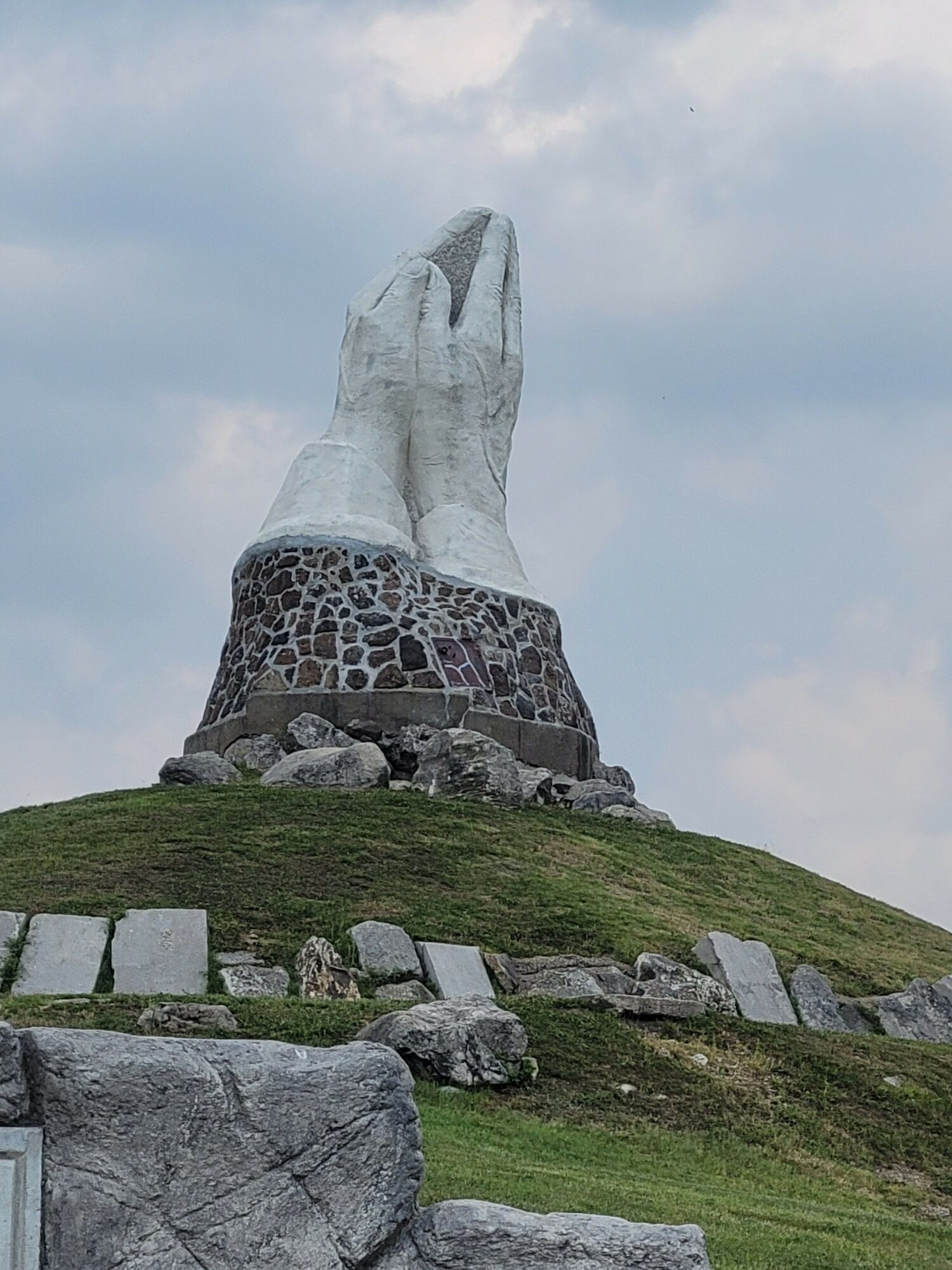 Large stone monument resembling a hand with fingers pointing upward, situated on a grassy hill with stones around base.