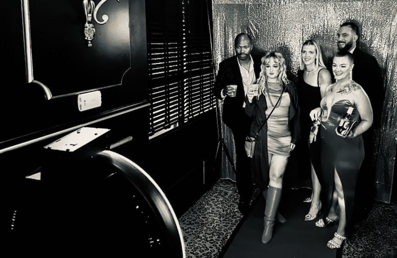 Group of five people standing together in a dimly lit room, some holding drinks and a helmet, with a textured wall behind.