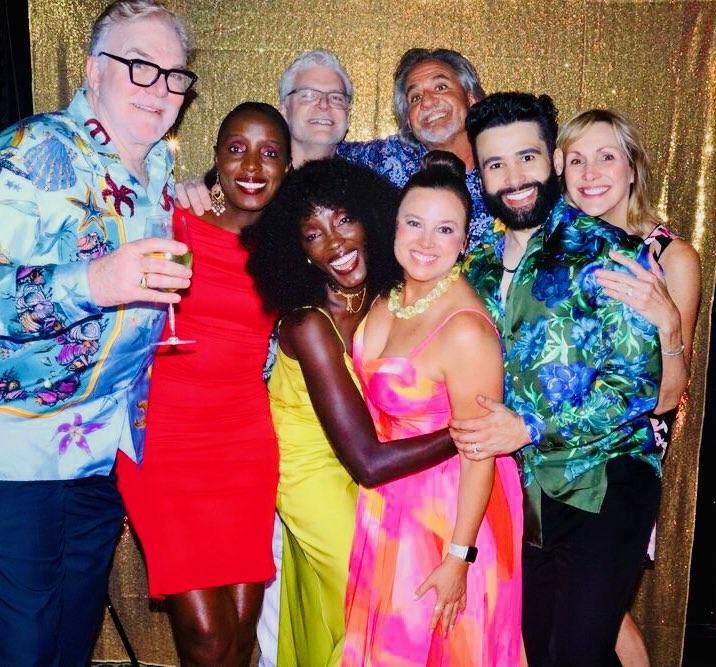 Group of nine diverse people smiling and posing together in front of a gold backdrop, dressed in colorful outfits.