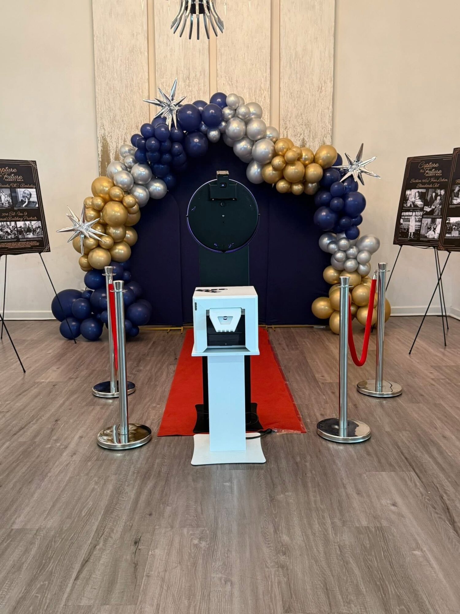 Balloon arch with blue, gold, and silver balloons over a doorway, red carpet, and stanchions with red ropes, in an indoor setting.