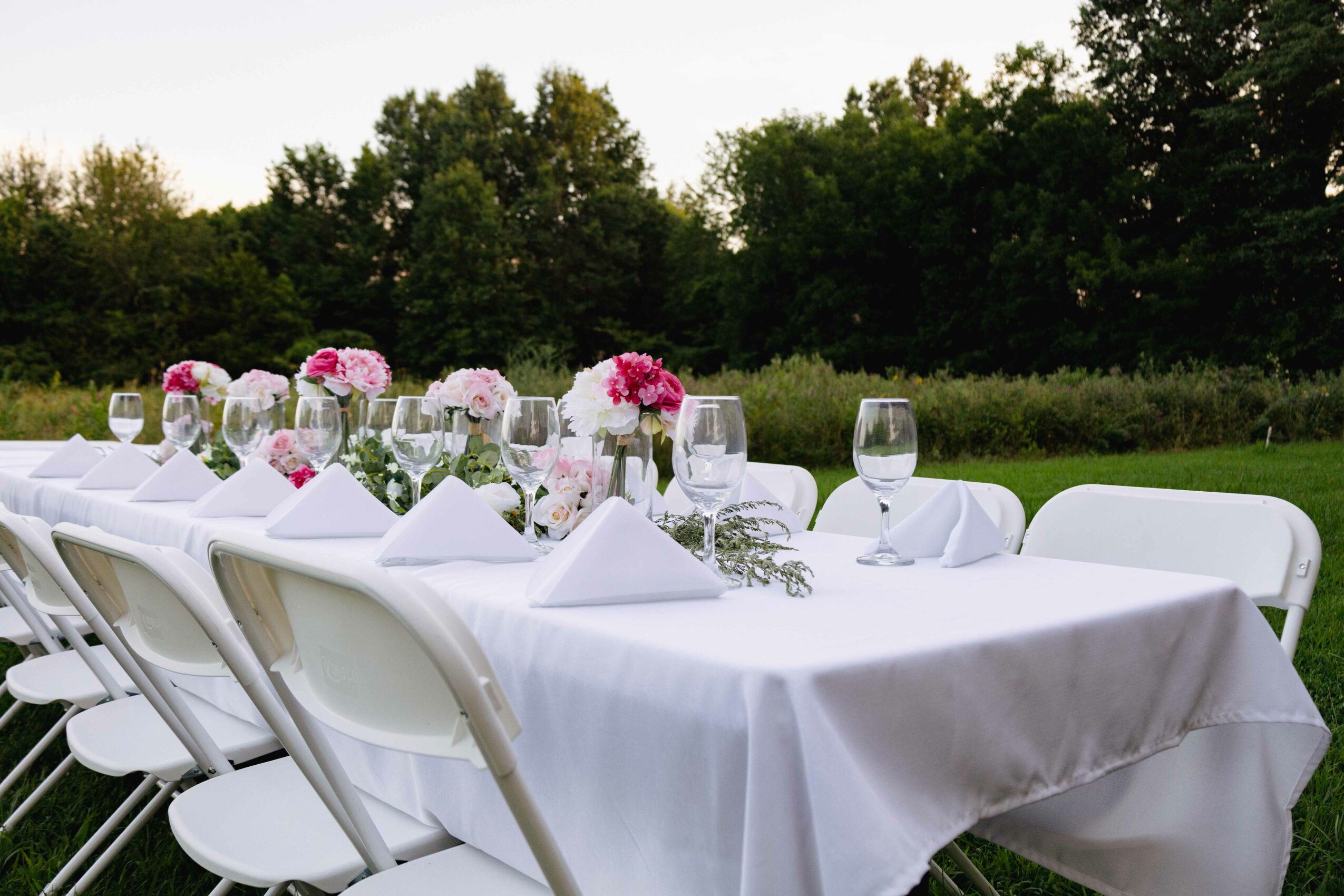 Long outdoor table set with white tablecloth, pink and white floral centerpieces, wine glasses, and white chairs, outdoors with trees.