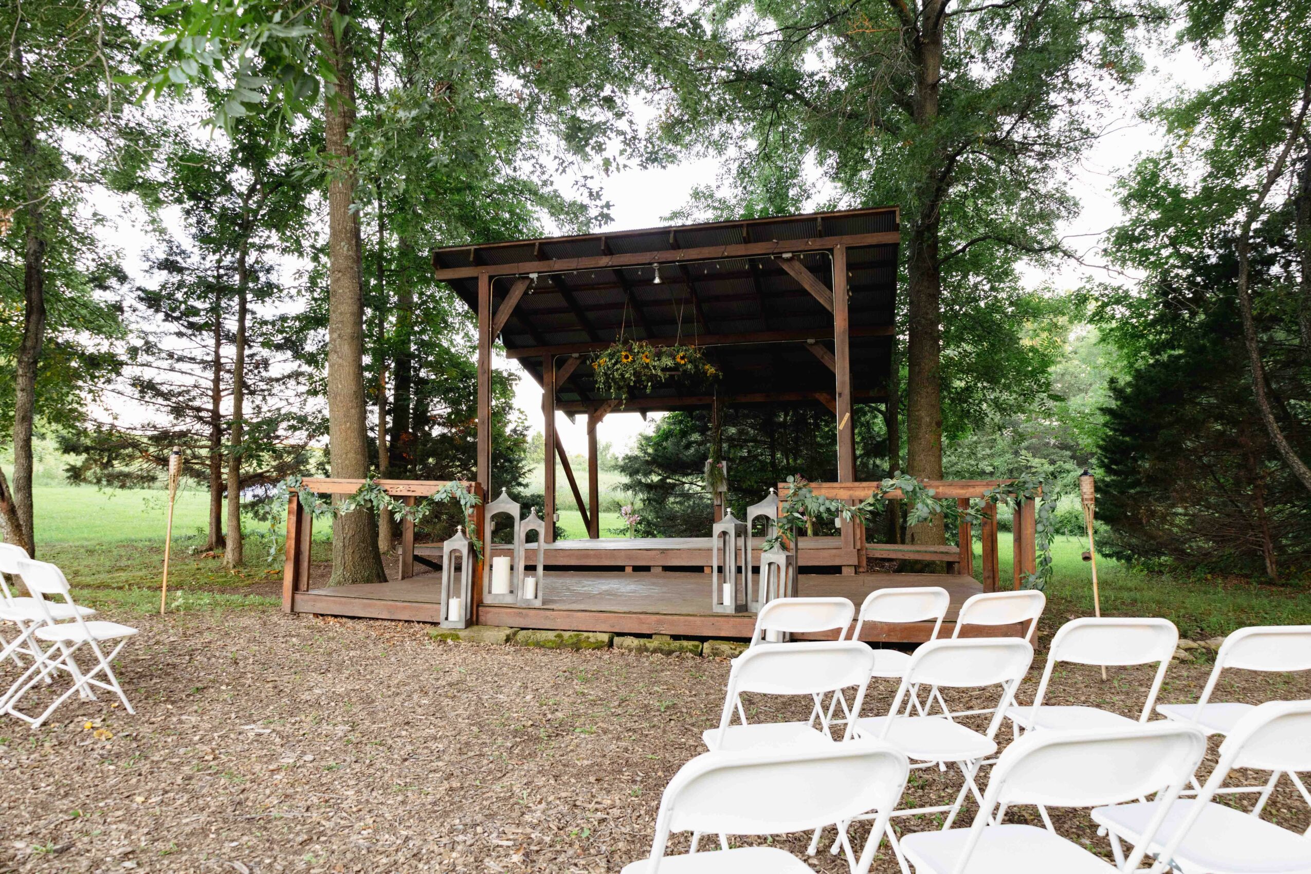 Outdoor wooden stage with a roof, surrounded by trees and empty white chairs on gravel ground.