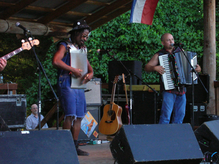 Two musicians perform on an outdoor stage with guitars and an accordion, surrounded by trees and a flag.