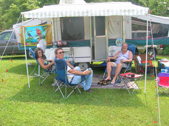 Four people sitting under a canopy in front of a camper trailer, with camping chairs and a colorful banner.