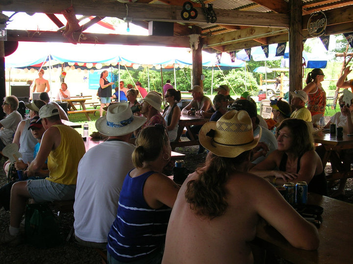 People sitting at tables under a wooden pavilion with colorful decorations, outdoors on a sunny day.