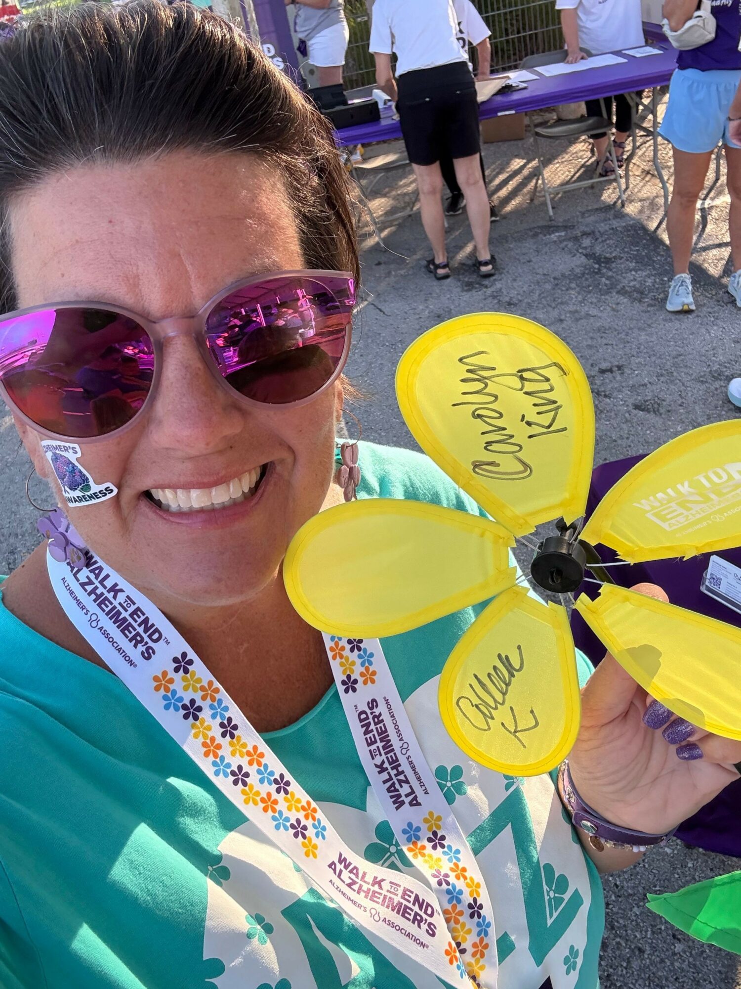 Woman smiling with sunglasses holding yellow and purple pinwheel, outdoor event with people in background.