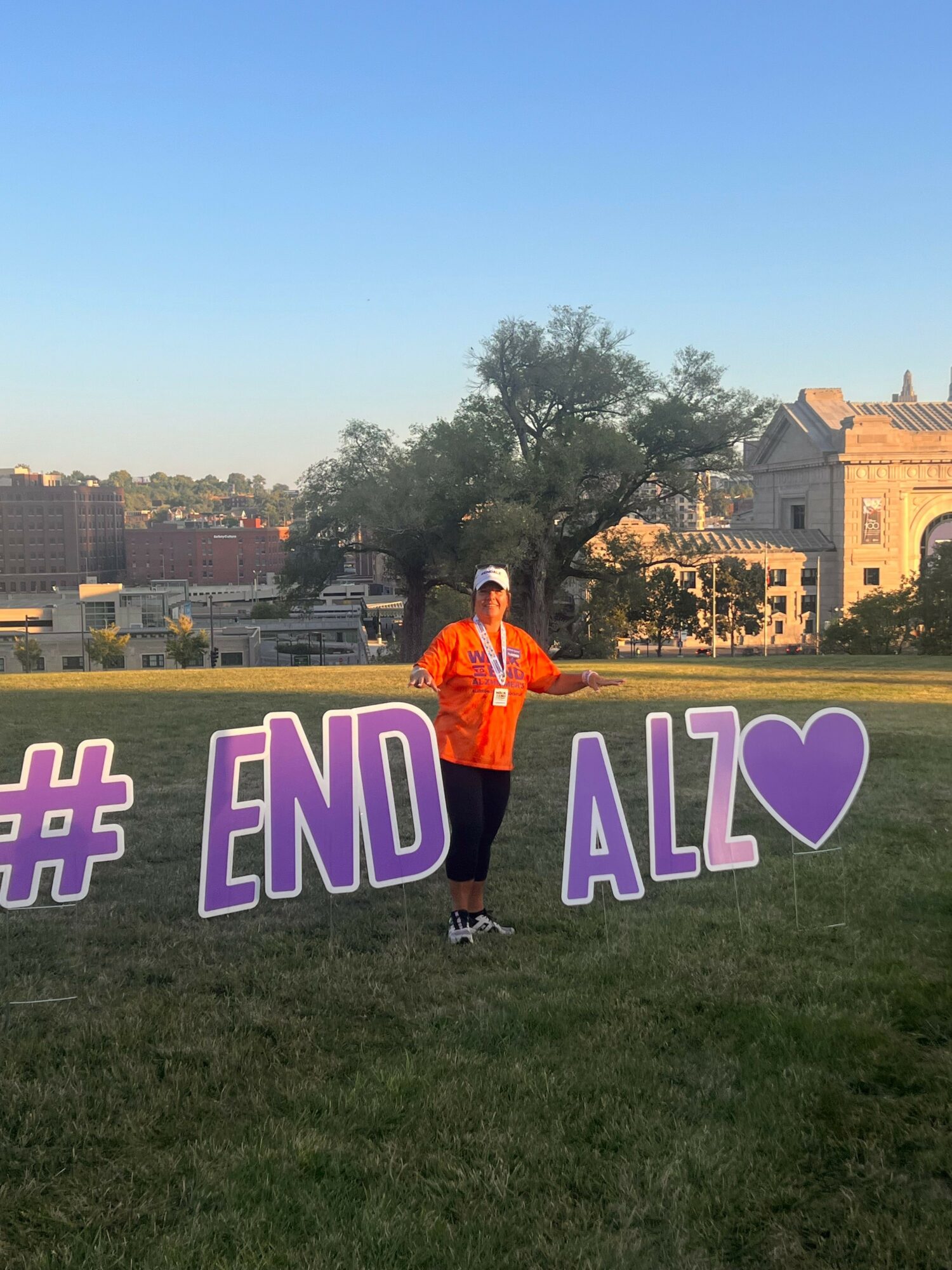 Person standing outdoors near large purple and white letters spelling '#ENDALZ' with a heart symbol, trees, and buildings in background.