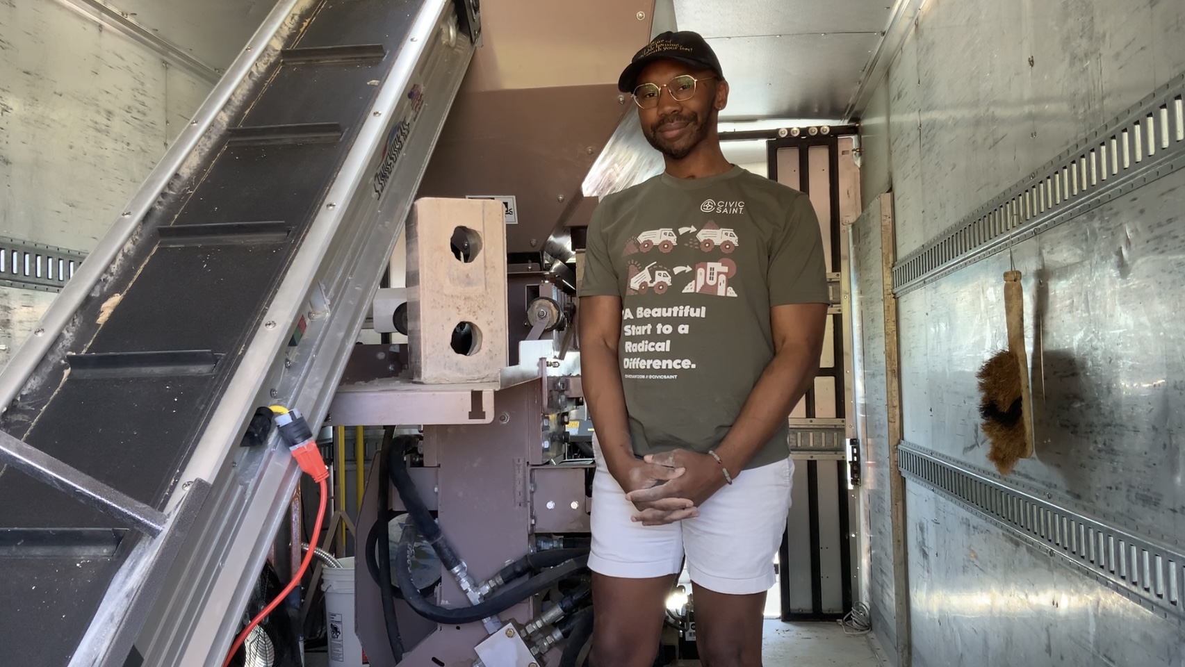 Person standing inside a laboratory or industrial setting, smiling, wearing glasses, a cap, a dark t-shirt, and white shorts.
