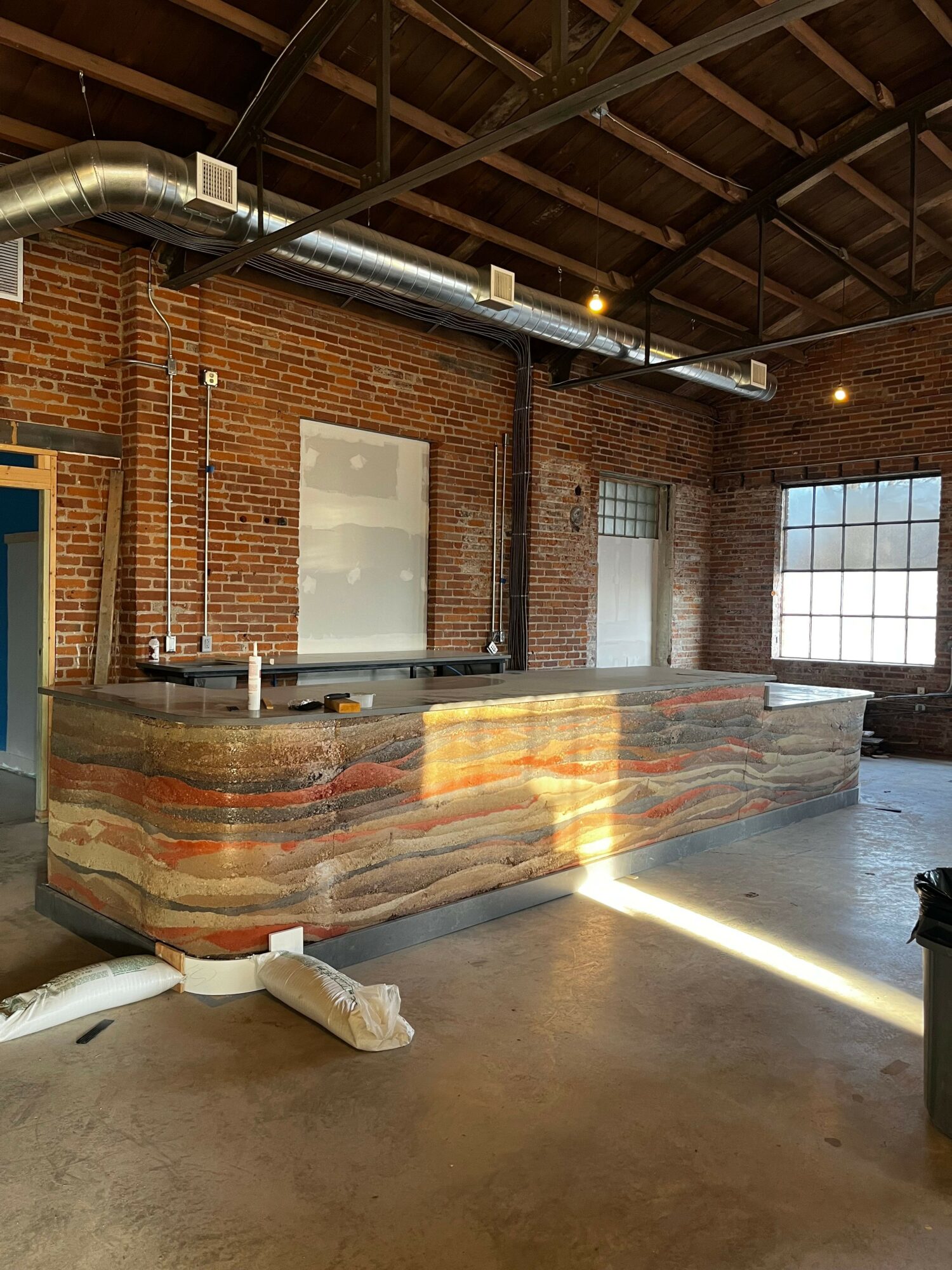 Brick wall with large window, industrial-style bar counter with colorful layered wood design, exposed ceiling beams, and ductwork.