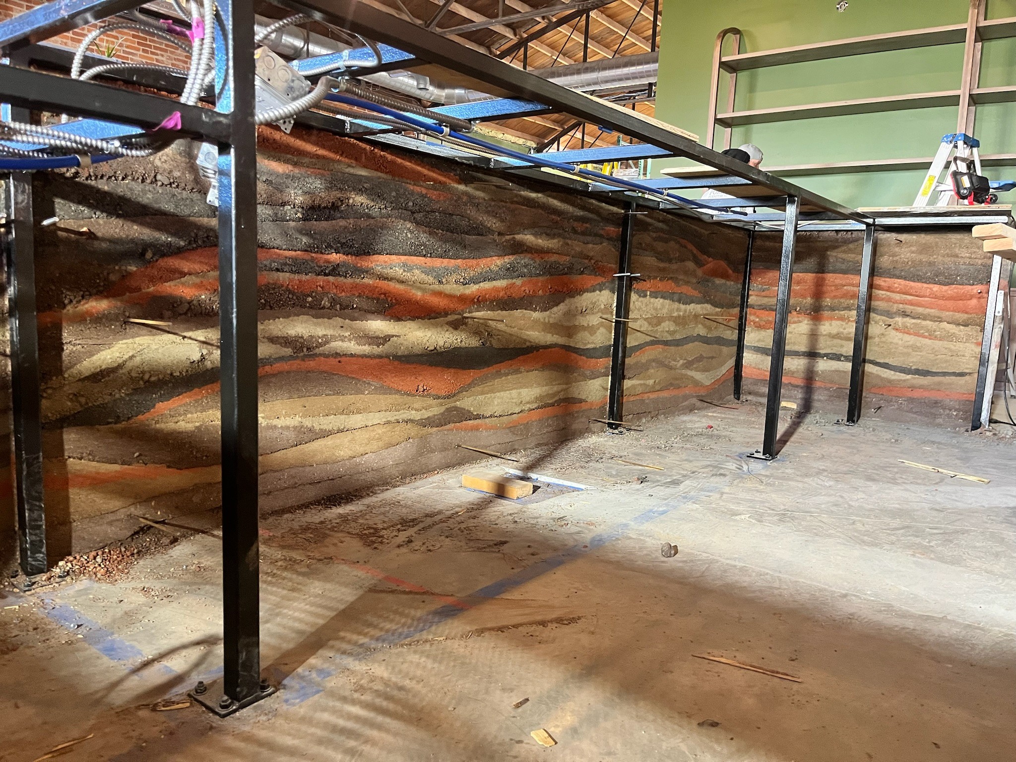 Metal framework with pipes and wires above a concrete floor, with a colorful layered wall in the background.