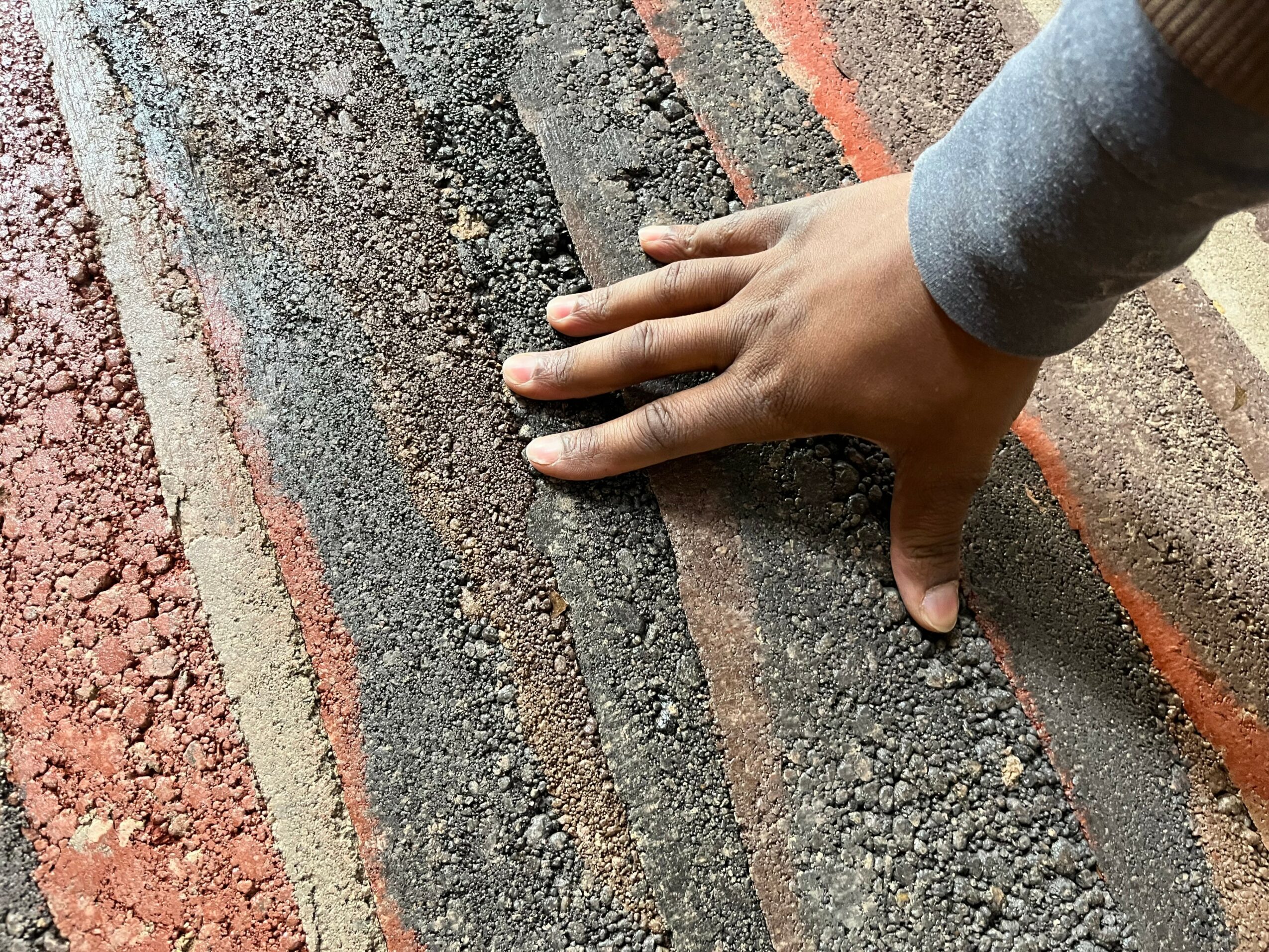 Person's hand touching textured asphalt surface with red and black stripes.