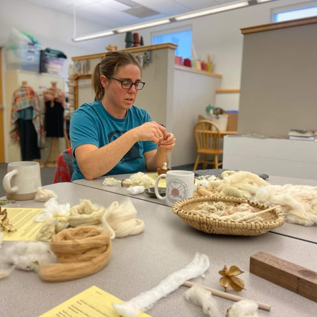 Woman with glasses working on textile project at table with yarn and tools in a craft room.