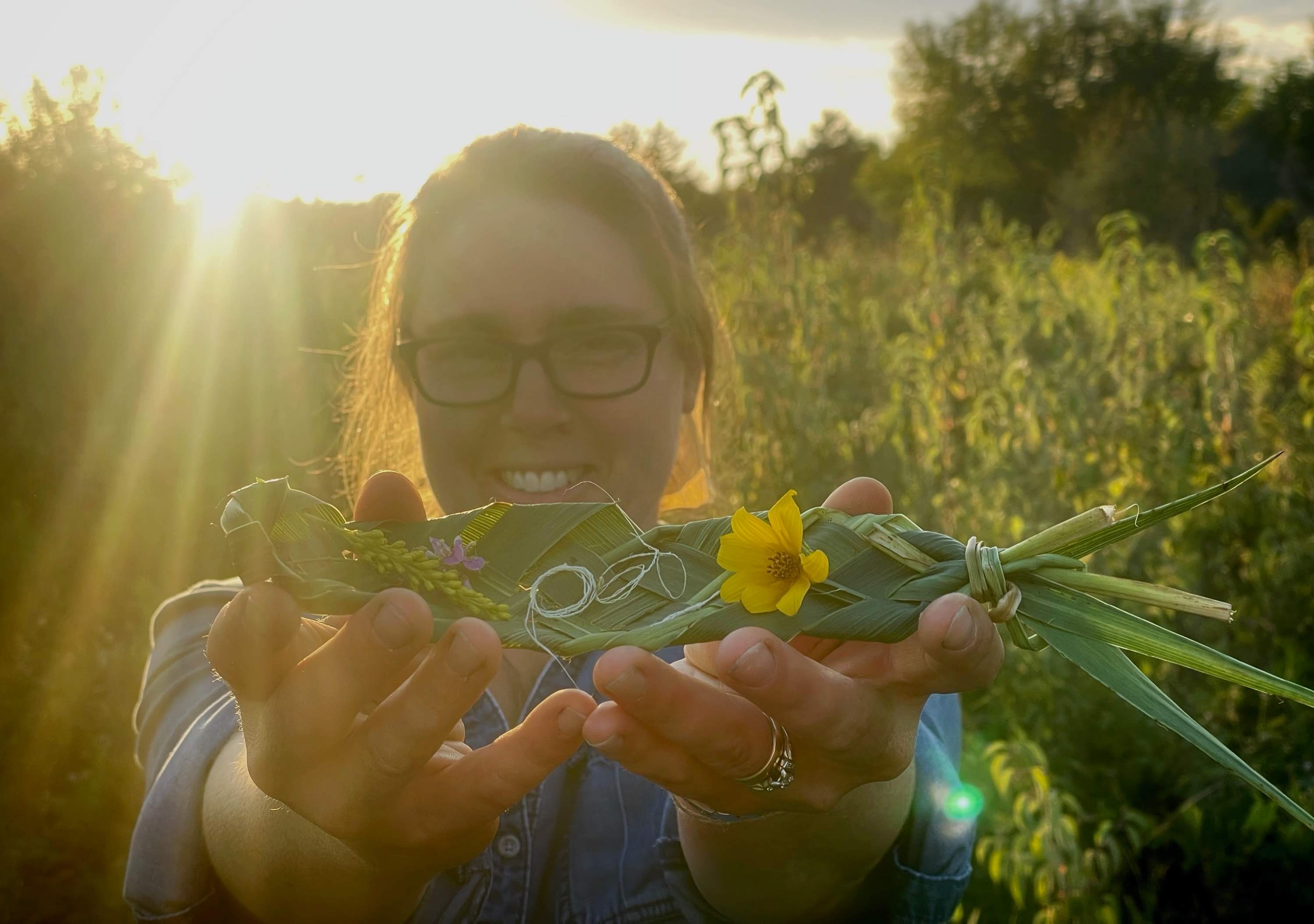 Person holding a decorated fish with flowers outdoors during sunset, smiling, wearing glasses.