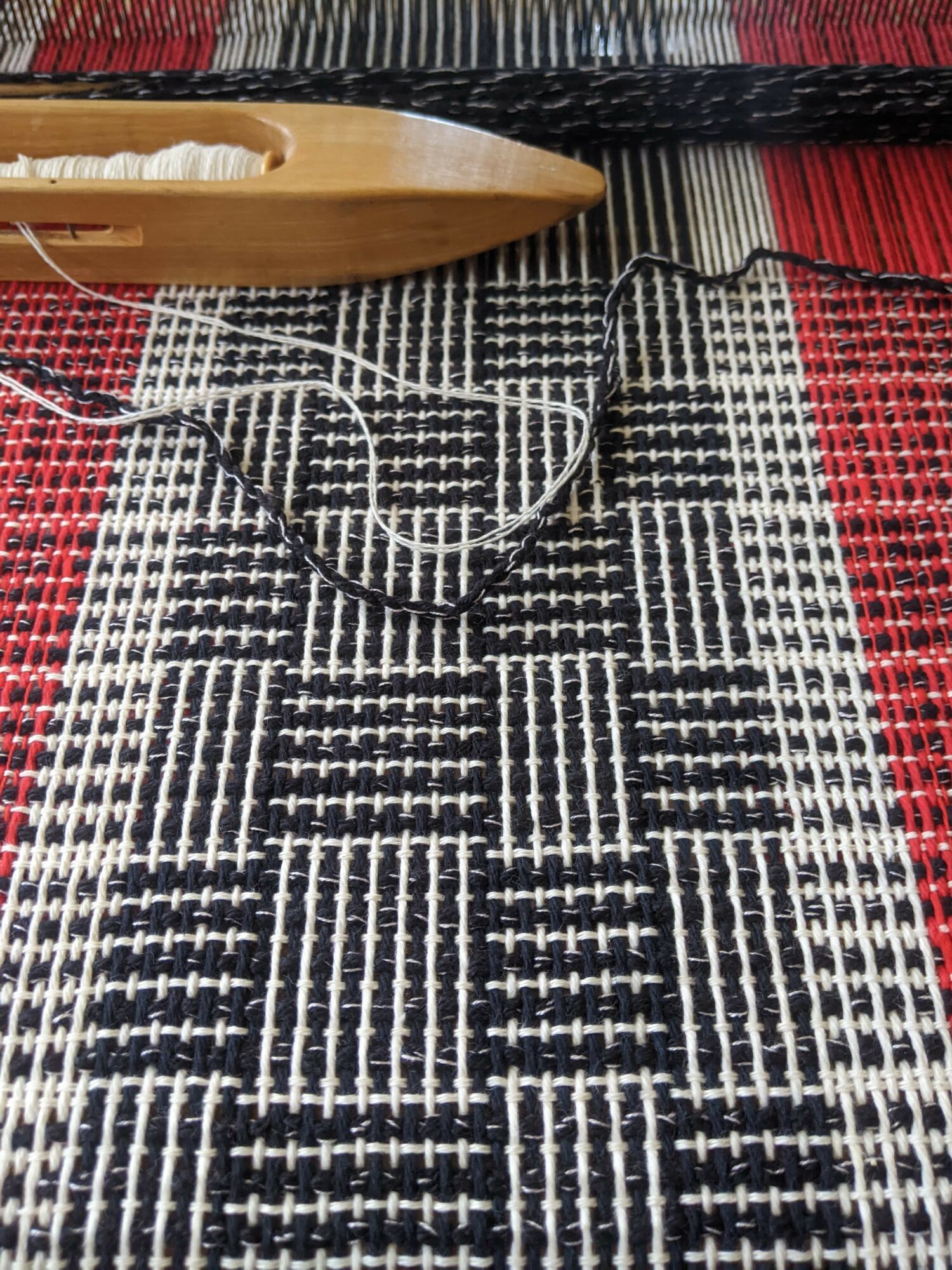 A wooden weaving shuttle and a black wire are placed on a black, white, and red woven textile.