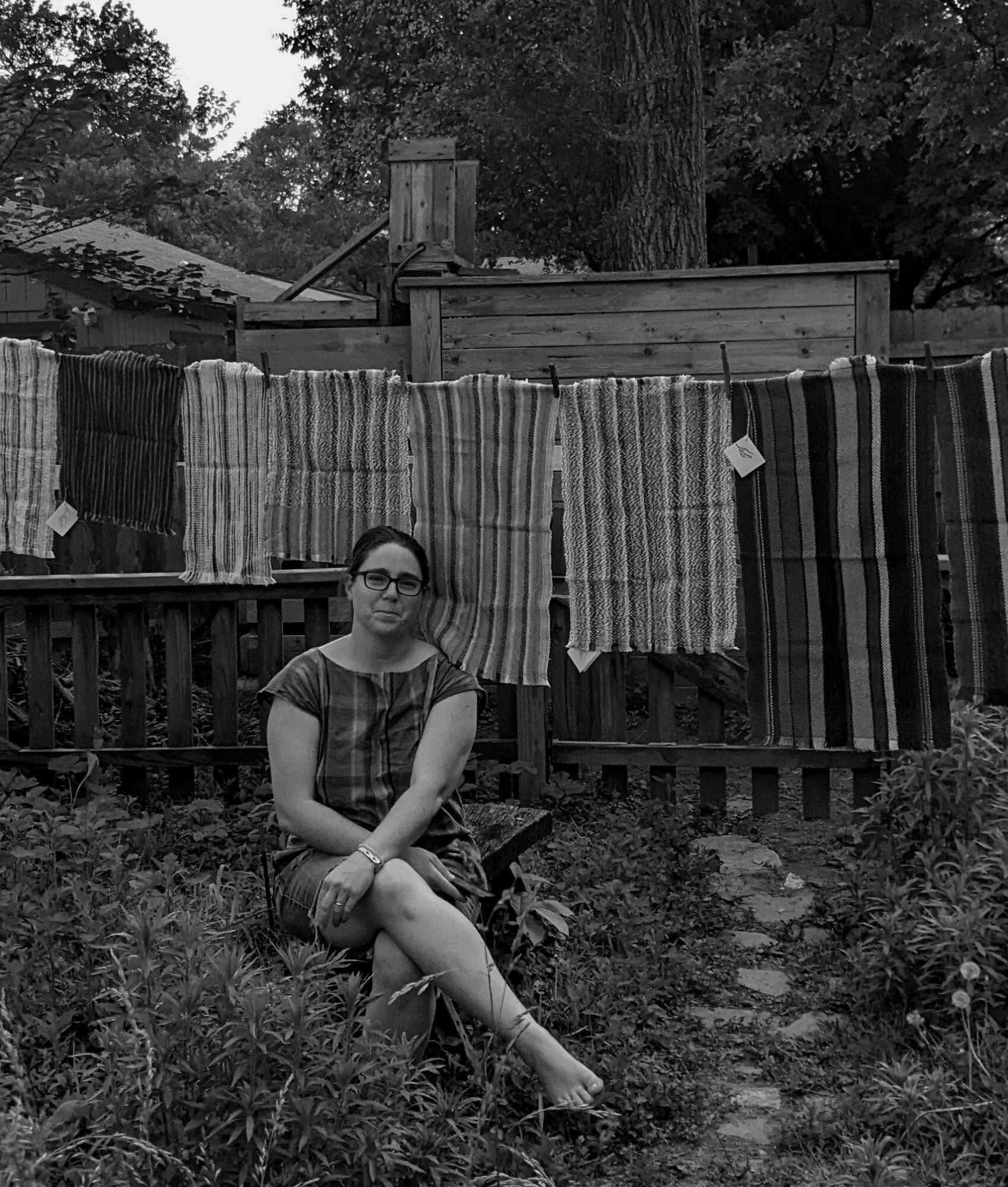 Woman sitting outdoors in front of a fence with hanging striped fabrics, surrounded by plants and trees.