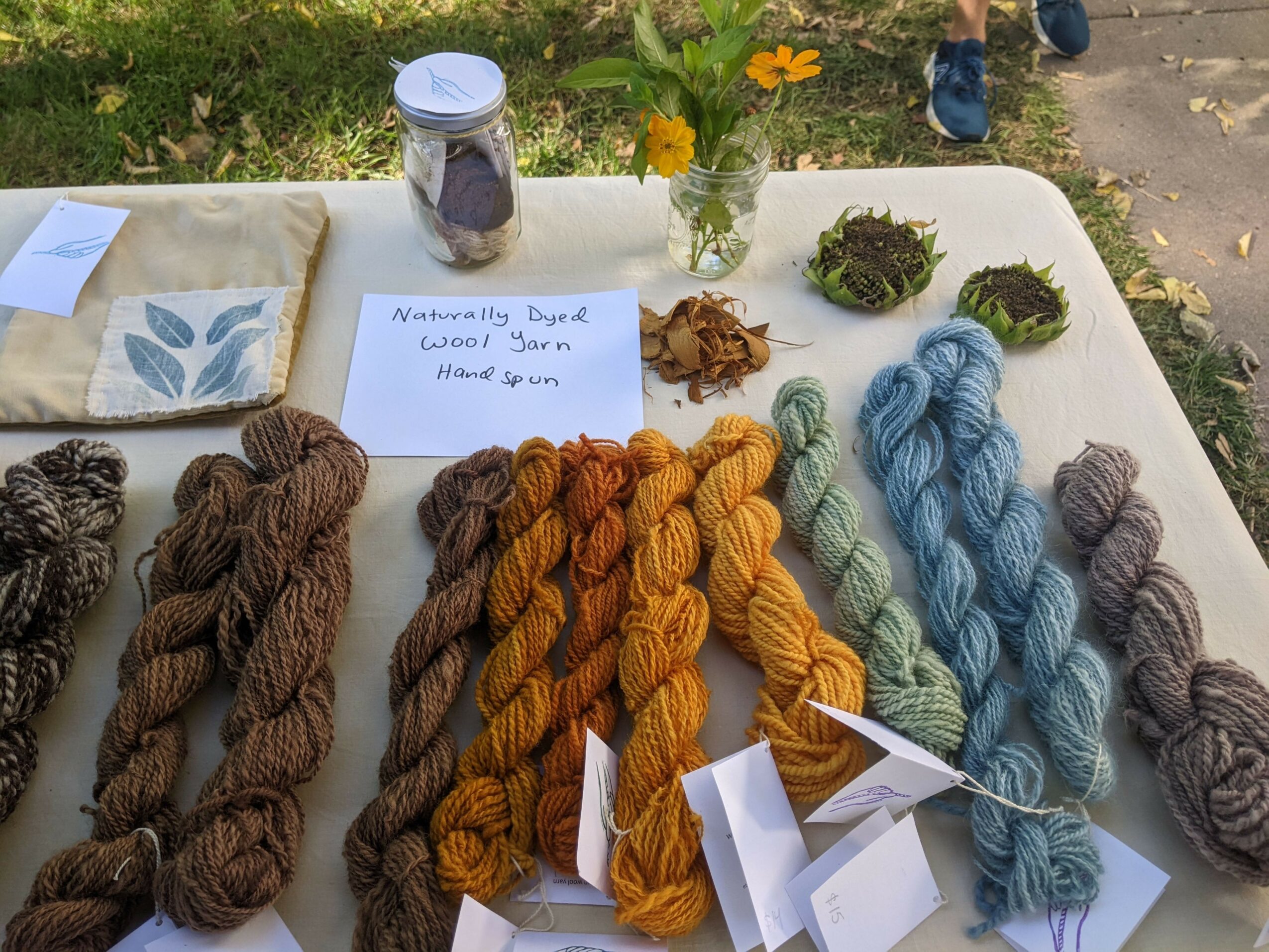 Display of colorful yarn skeins, flowers, and a jar on a white table outdoors, with a handwritten note.