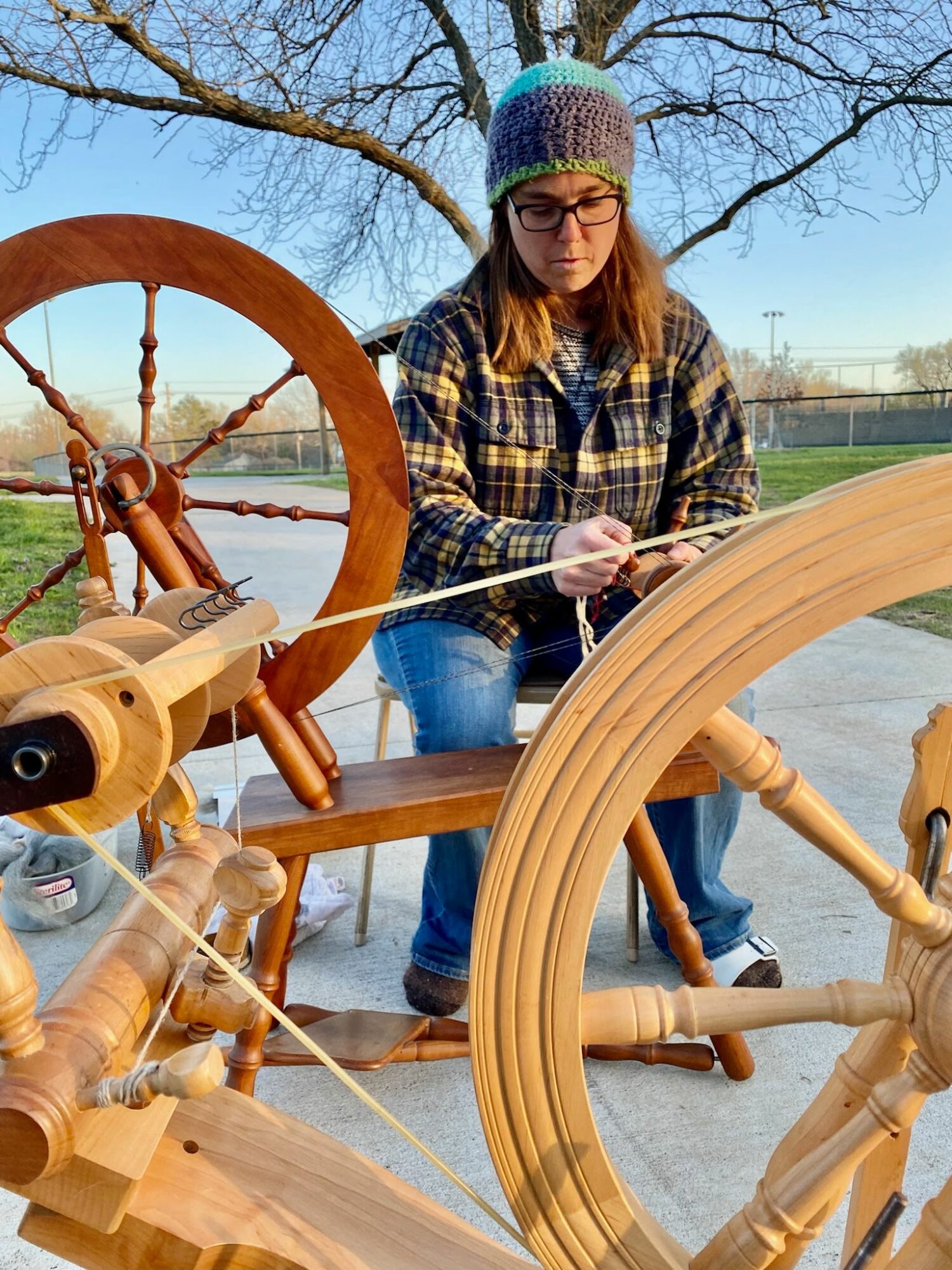 Young woman wearing glasses, a colorful knit hat, and plaid shirt, working on a large wooden spinning wheel outdoors.