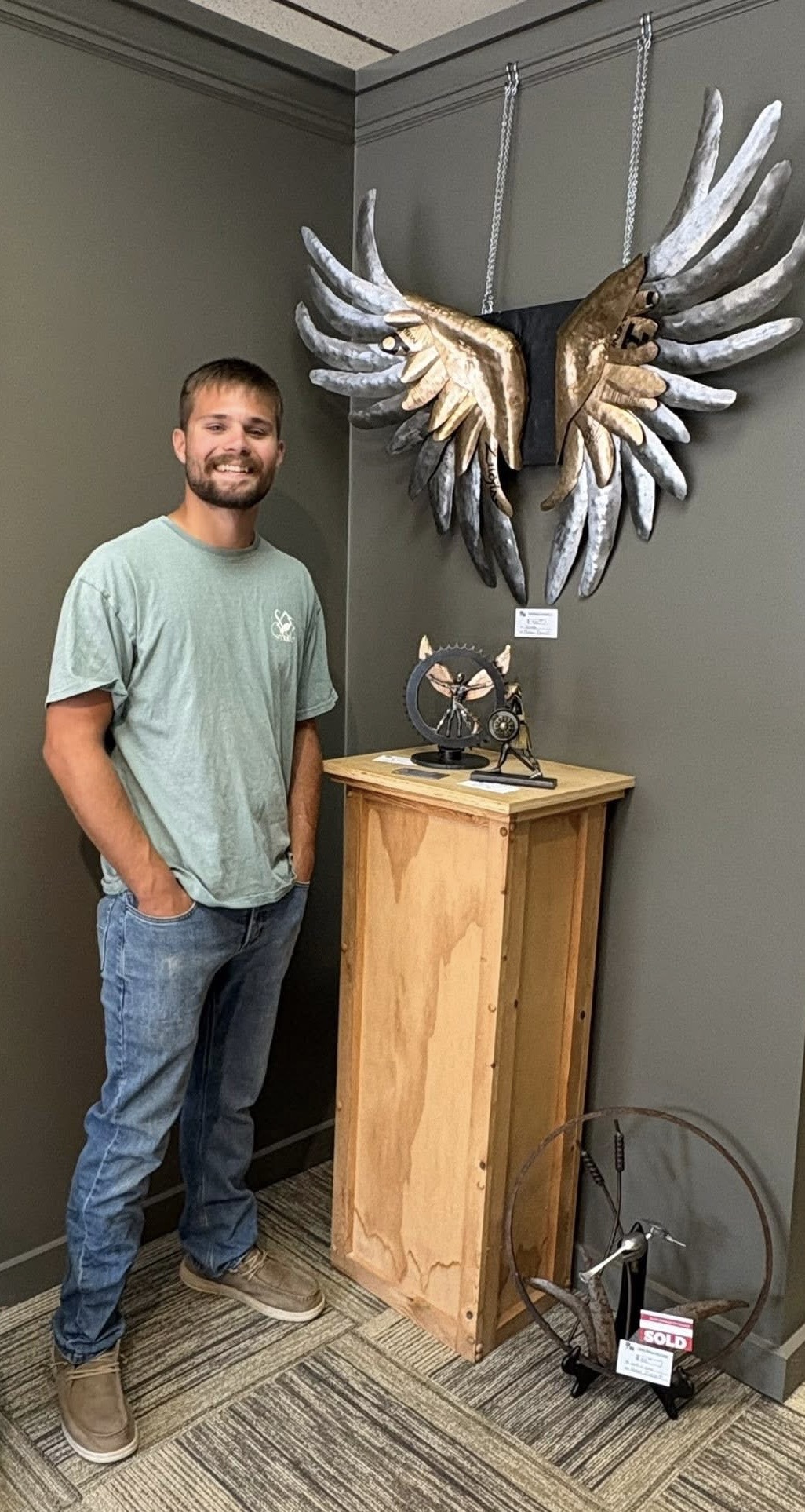 Man with beard and casual clothing standing next to a wooden pedestal with a decorative owl sculpture and a large hanging art piece with multiple hands.
