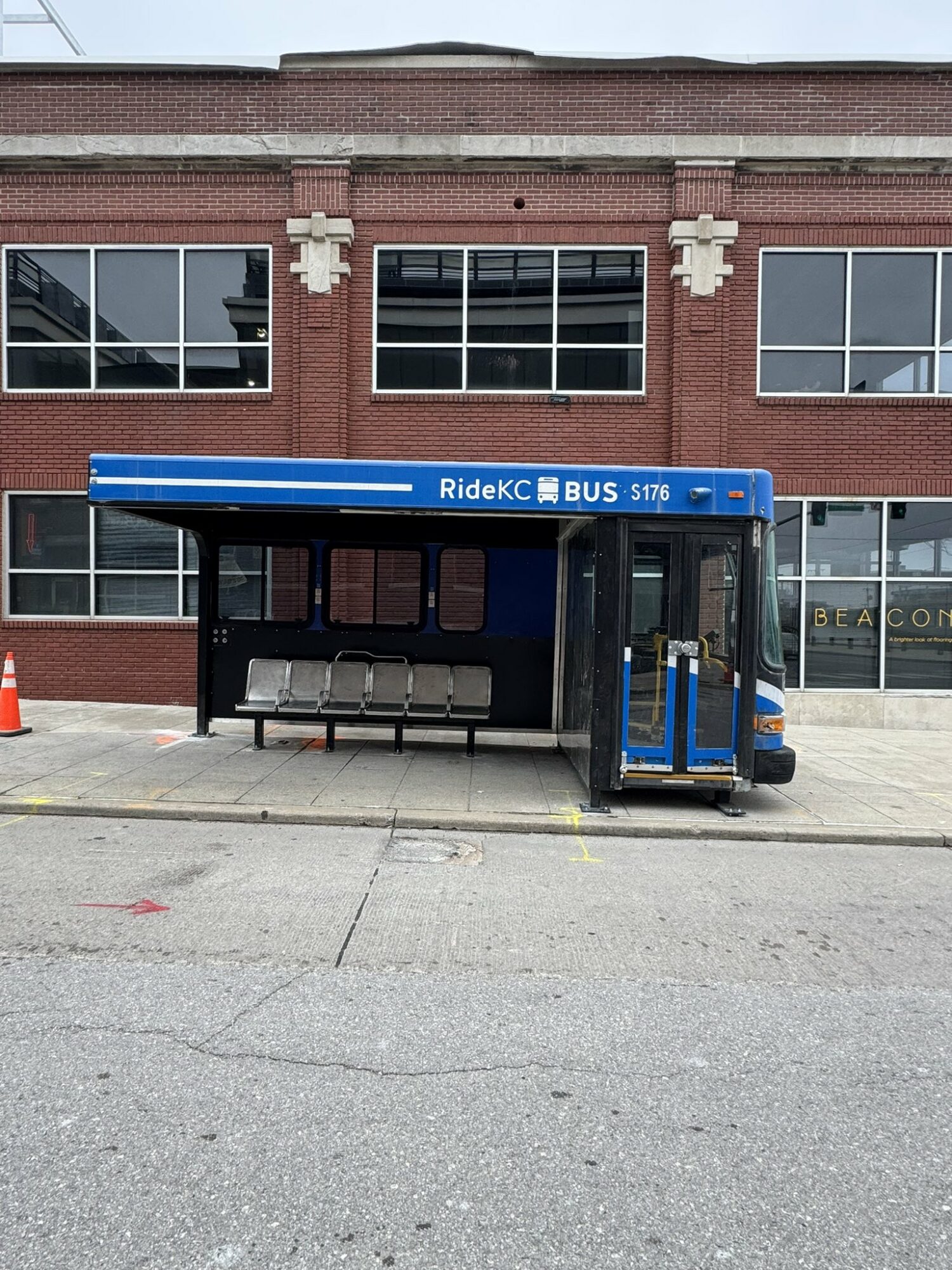 Bus shelter with blue roof and seating, in front of a brick building with large windows.