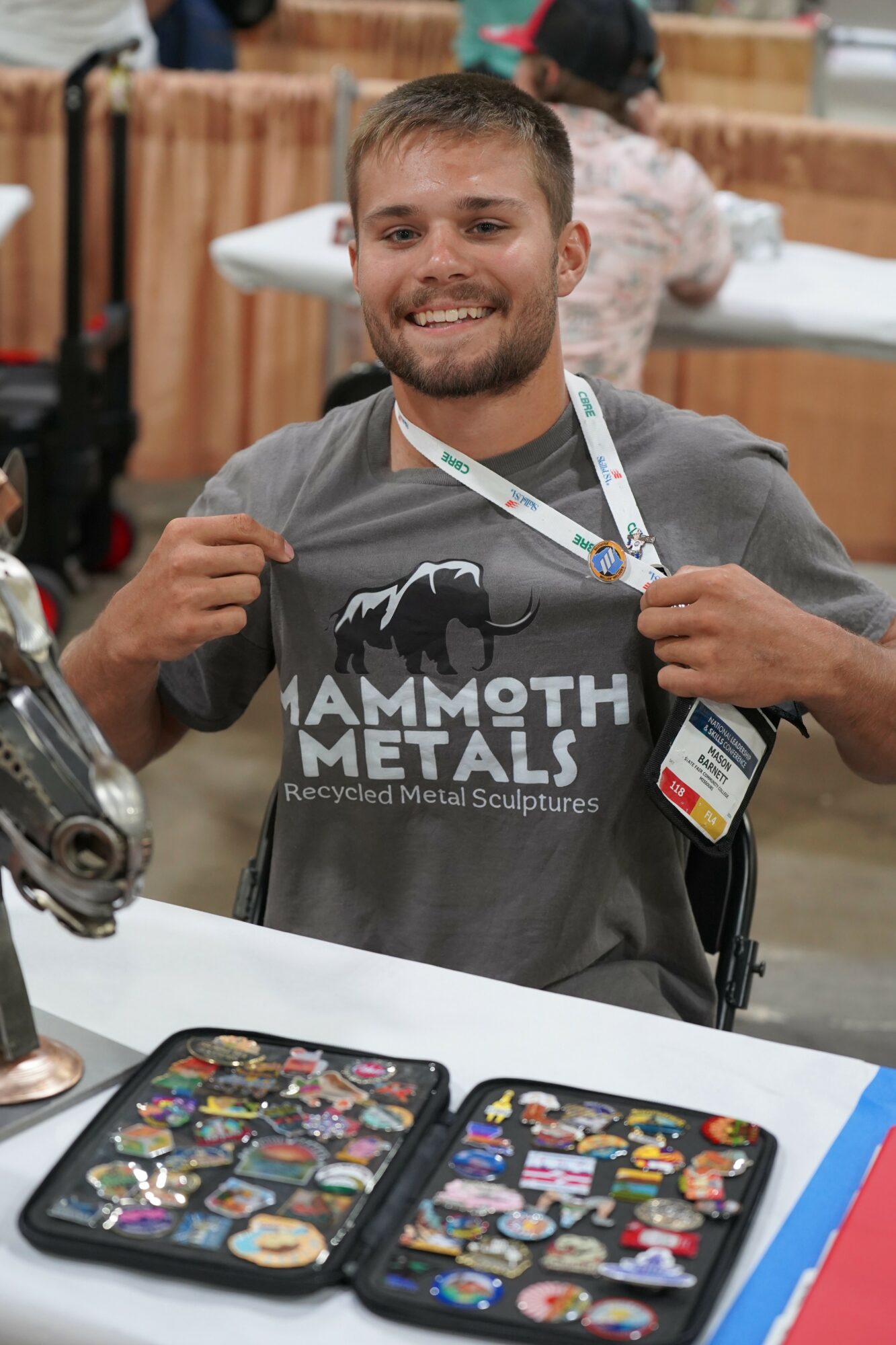 Young man with short hair and beard smiling, holding a gray T-shirt with a mammoth logo, at an event table.