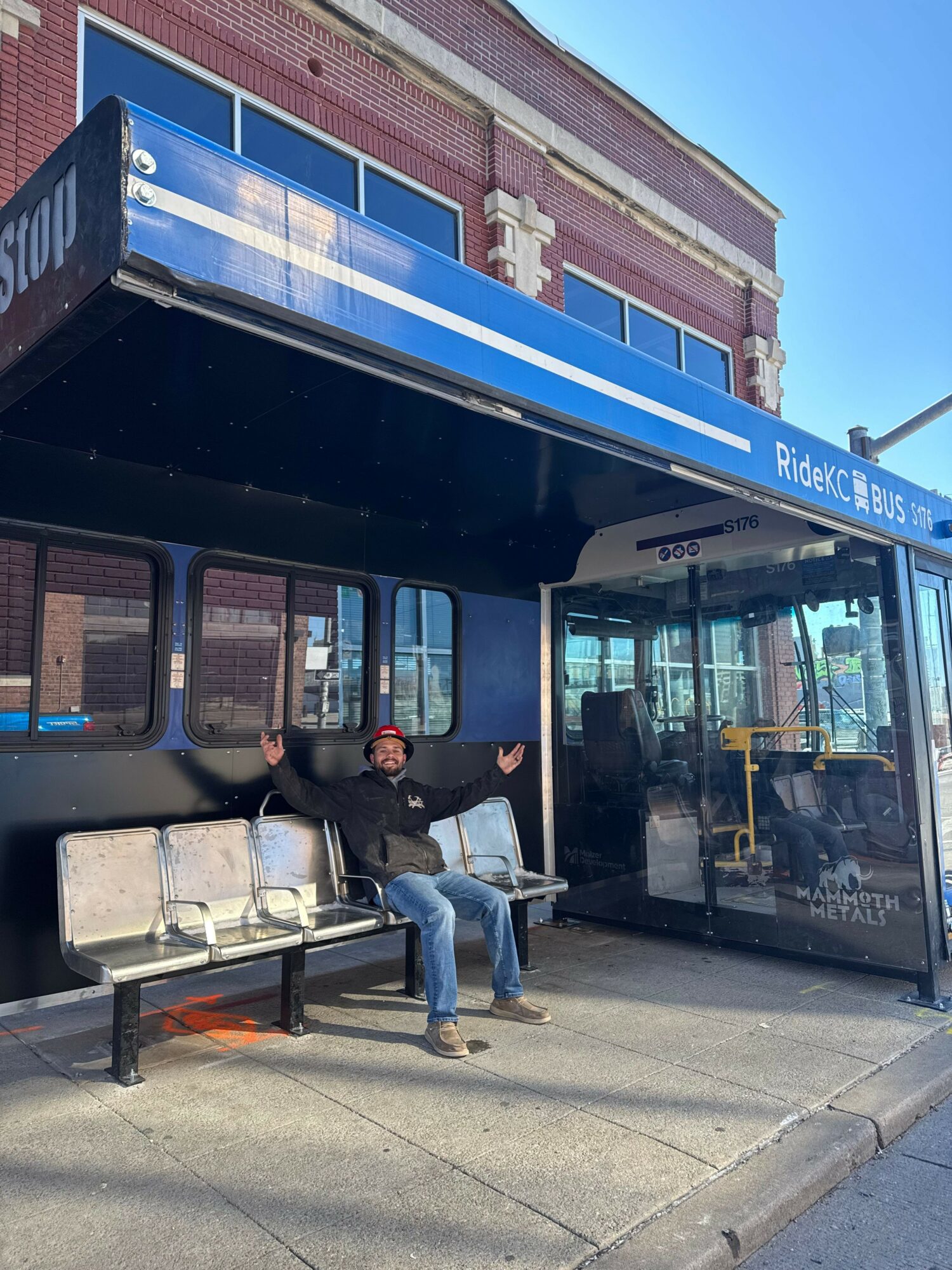 Person sitting on bench at bus stop with arms outstretched, building and bus shelter in background.