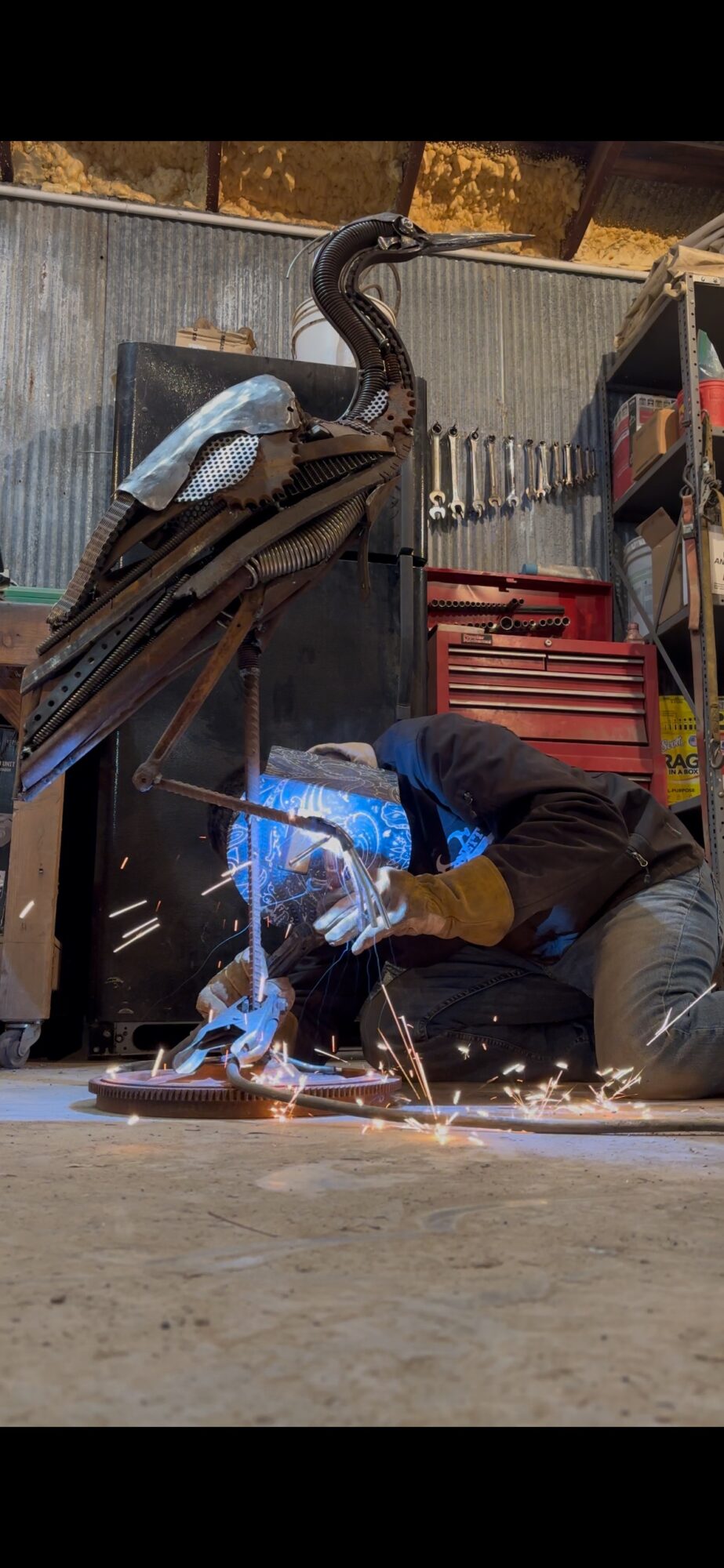 Person welding metal outdoors with sparks flying, tools and equipment nearby, in a workshop setting.