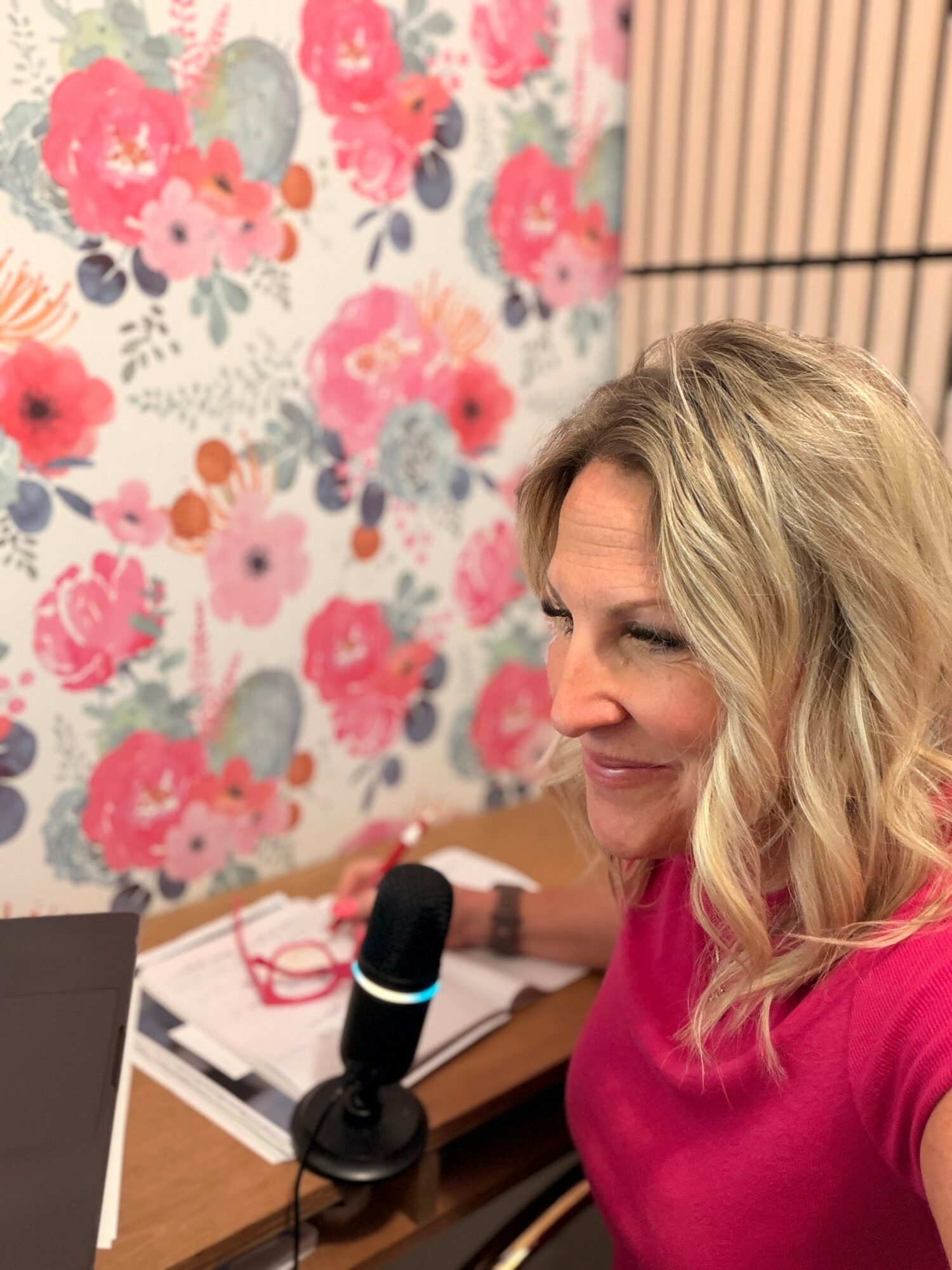 Woman with blonde hair smiling, wearing a pink shirt, sitting at a desk with a microphone, floral wallpaper background.