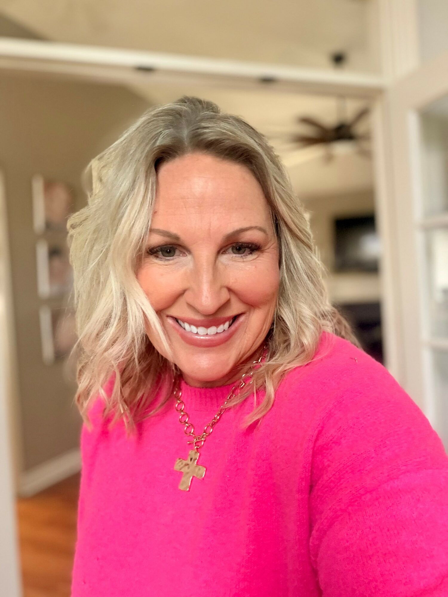 Smiling woman with blonde hair wearing a pink top and gold cross necklace indoors.