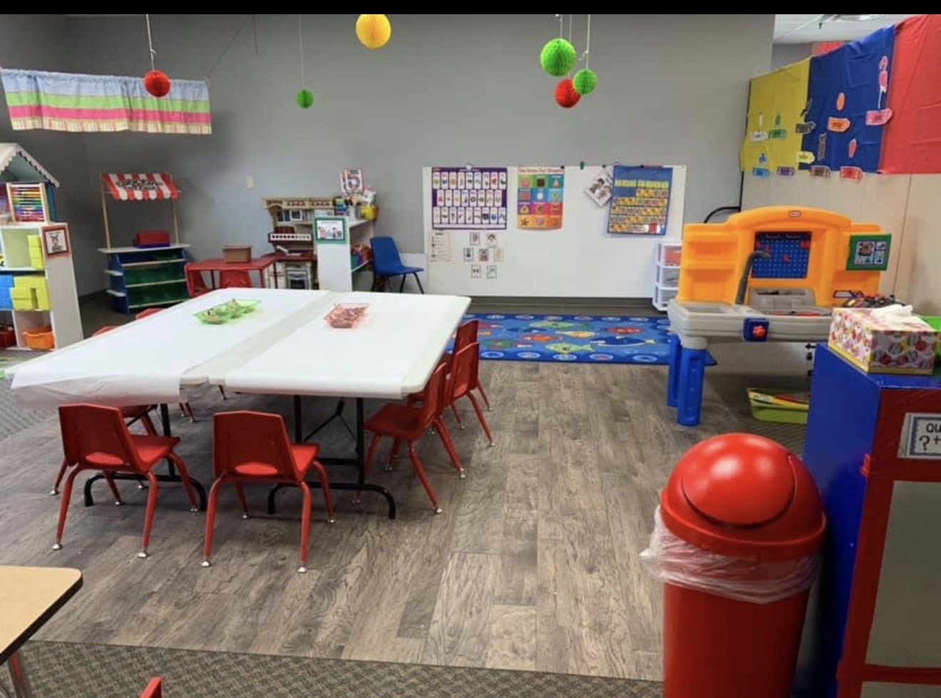 Classroom with tables, chairs, colorful decorations, and educational materials, including a play area and storage units.