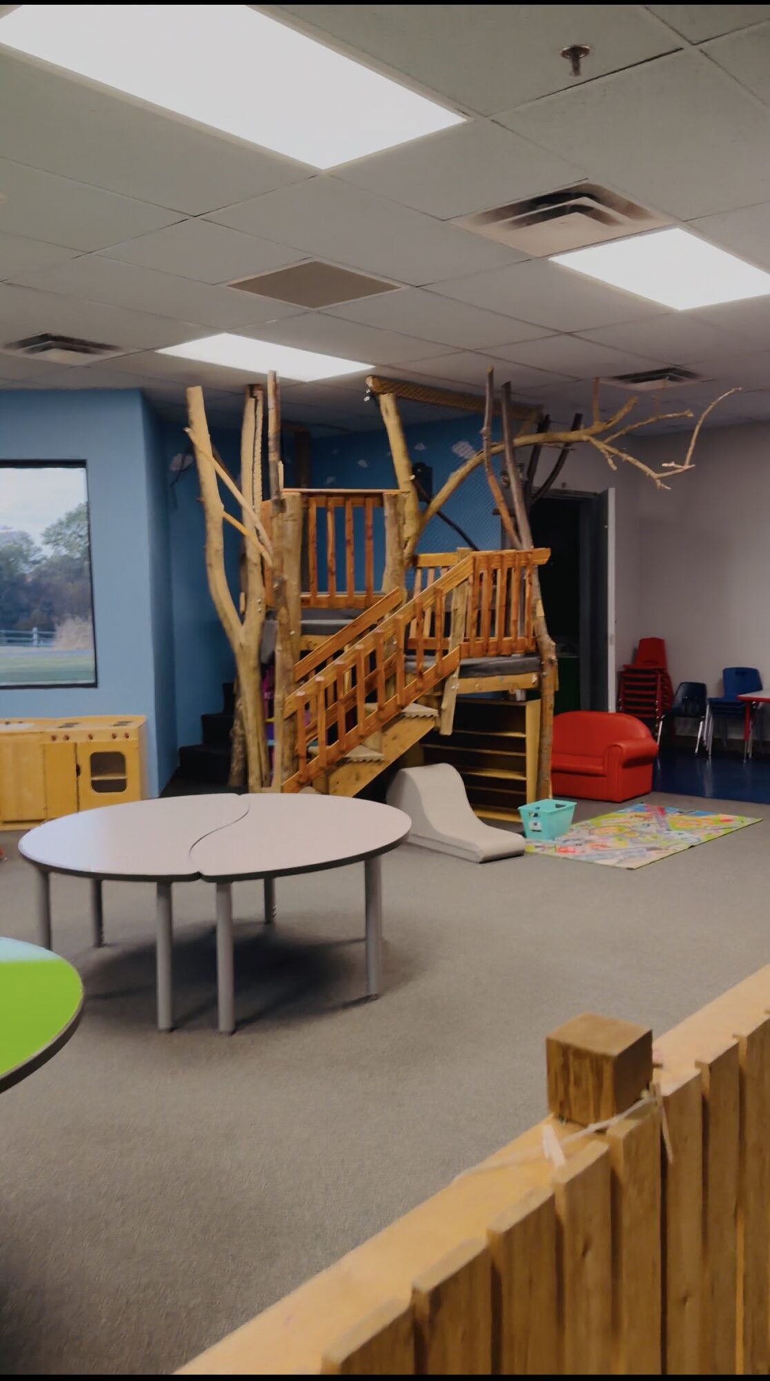Indoor play area with a treehouse structure, slide, tables, and chairs, enclosed by a wooden fence.