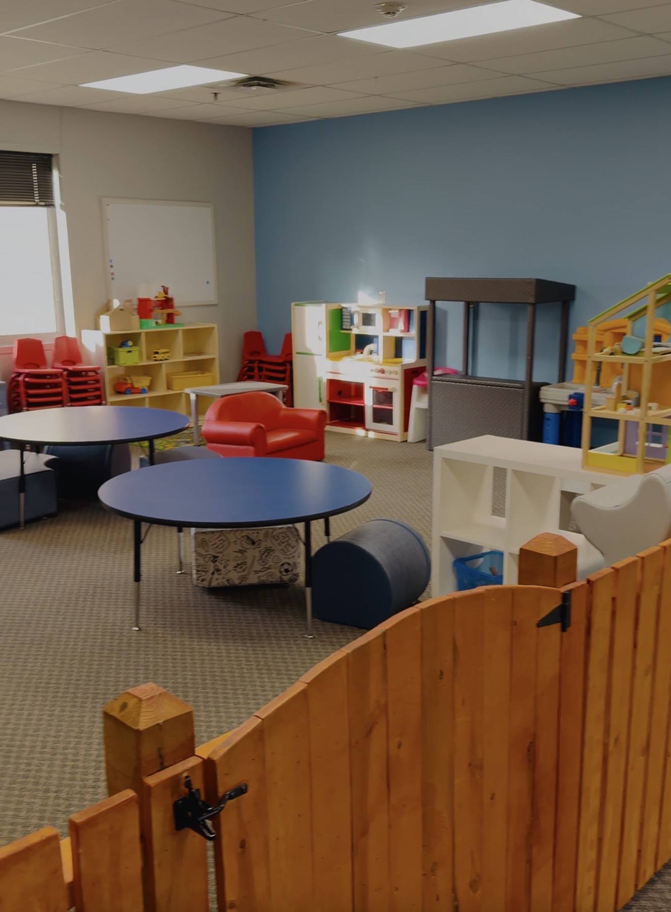 Children's playroom with colorful furniture, tables, and storage units, enclosed by a wooden fence, with a blue wall and ceiling lights.