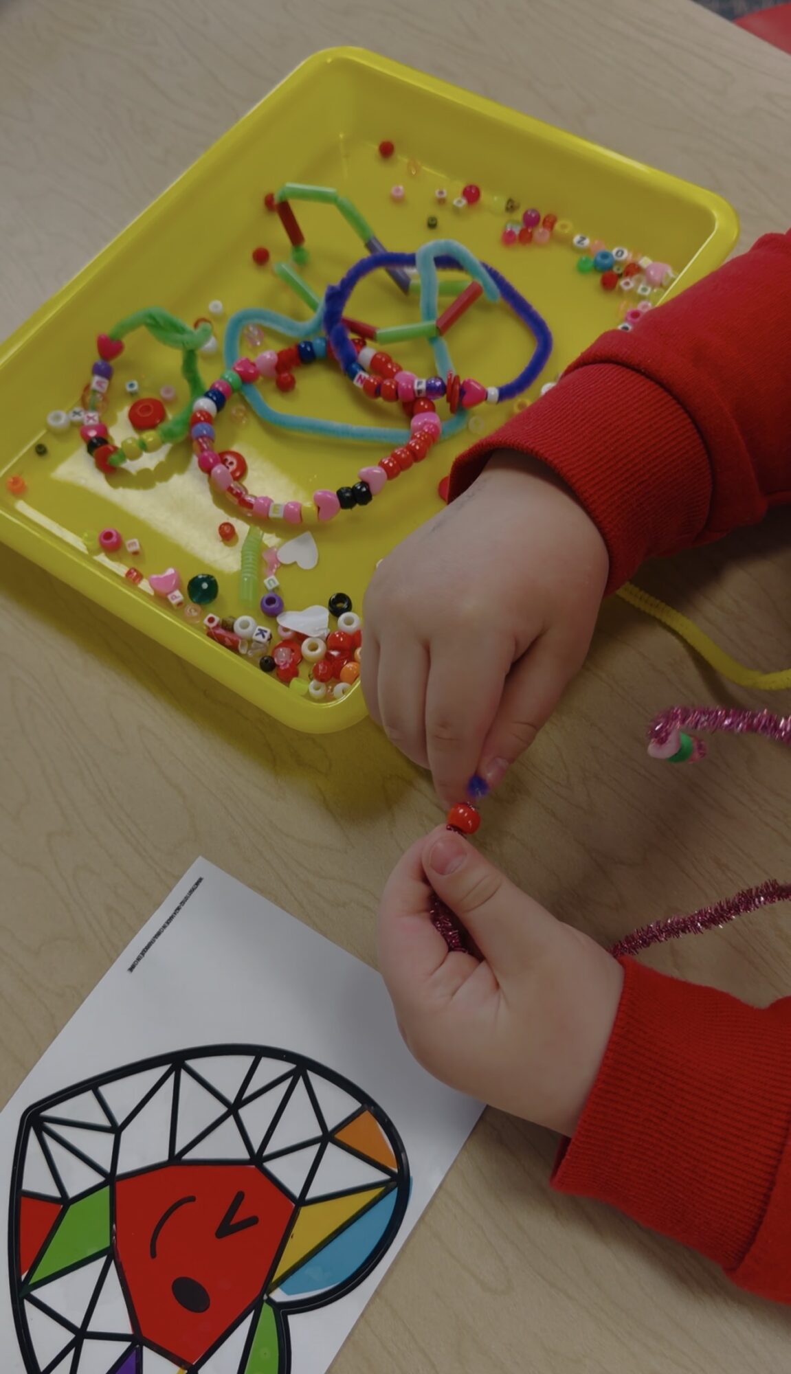 Child's hand stringing beads on a bracelet in a yellow tray with colorful beads and jewelry pieces.
