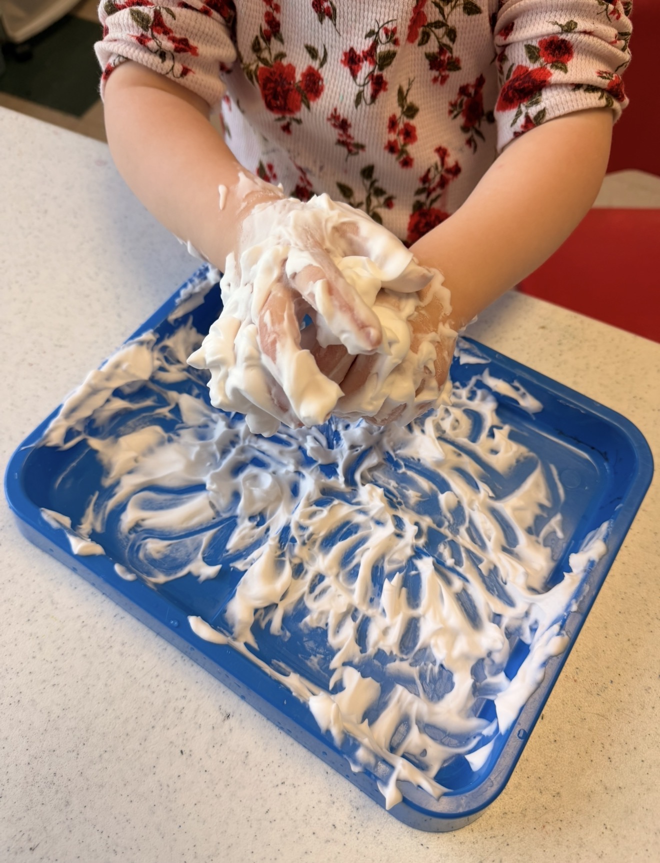Child's hands mixing white substance in a blue tray on a white surface, wearing a floral-patterned shirt.