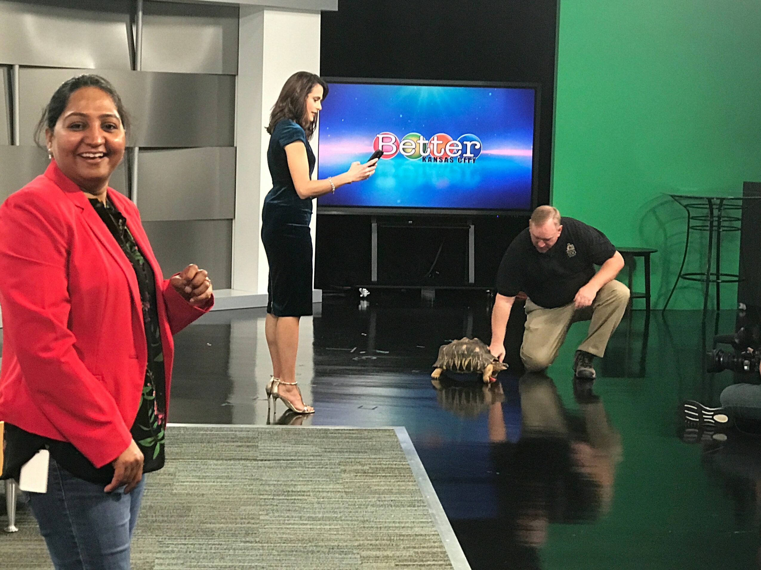 Studio with three people, one woman smiling in foreground, two others setting up equipment, green and black backgrounds, TV screen displaying 'Batter'.