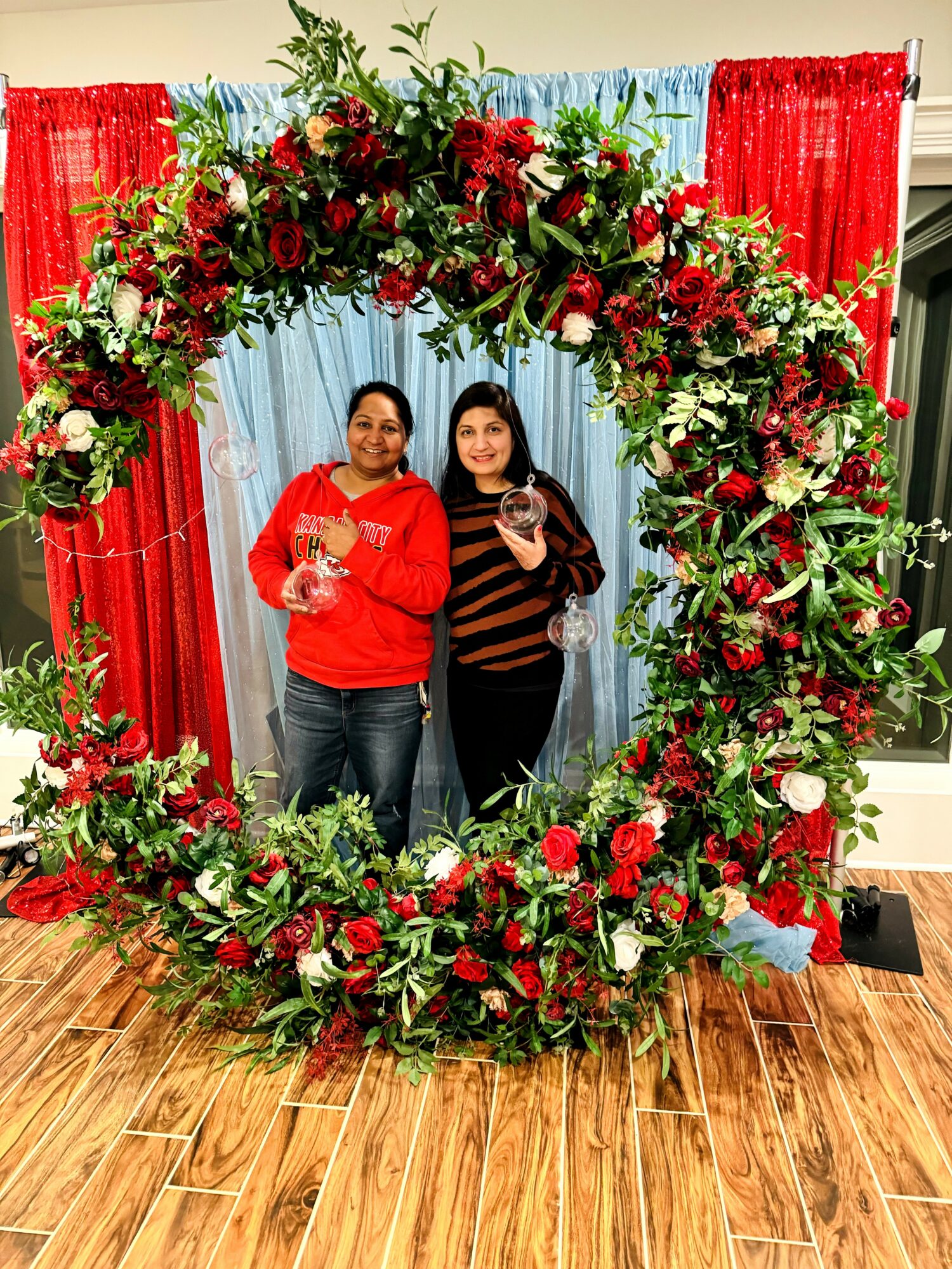 Two women stand inside a large floral heart-shaped frame with red, white, and green flowers, in front of colorful curtains.