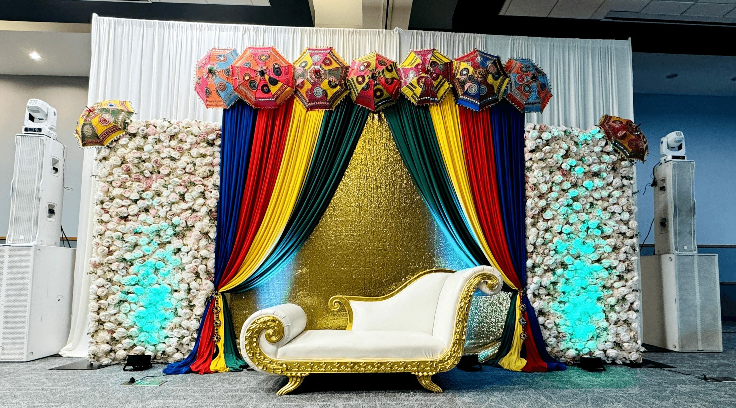 Decorative backdrop with colorful drapes, balloons, and floral arrangements, featuring a white and gold ornate sofa in front.