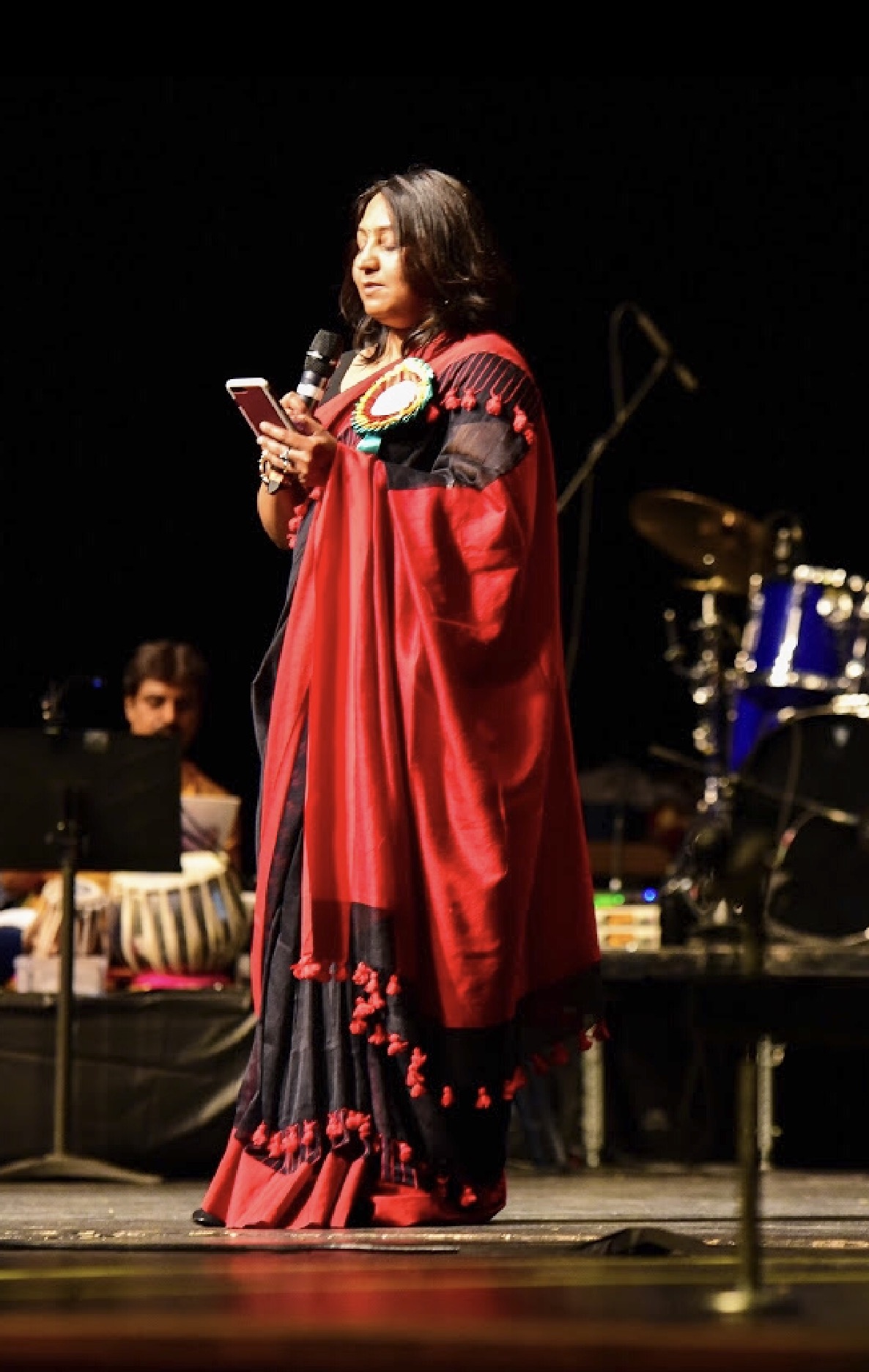 Woman in a red and black traditional dress holding a phone on stage with musical instruments in background.