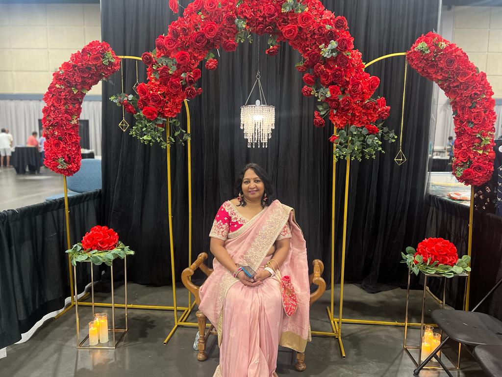 Woman in pink saree sitting on a chair with floral decorations and black backdrop behind her.