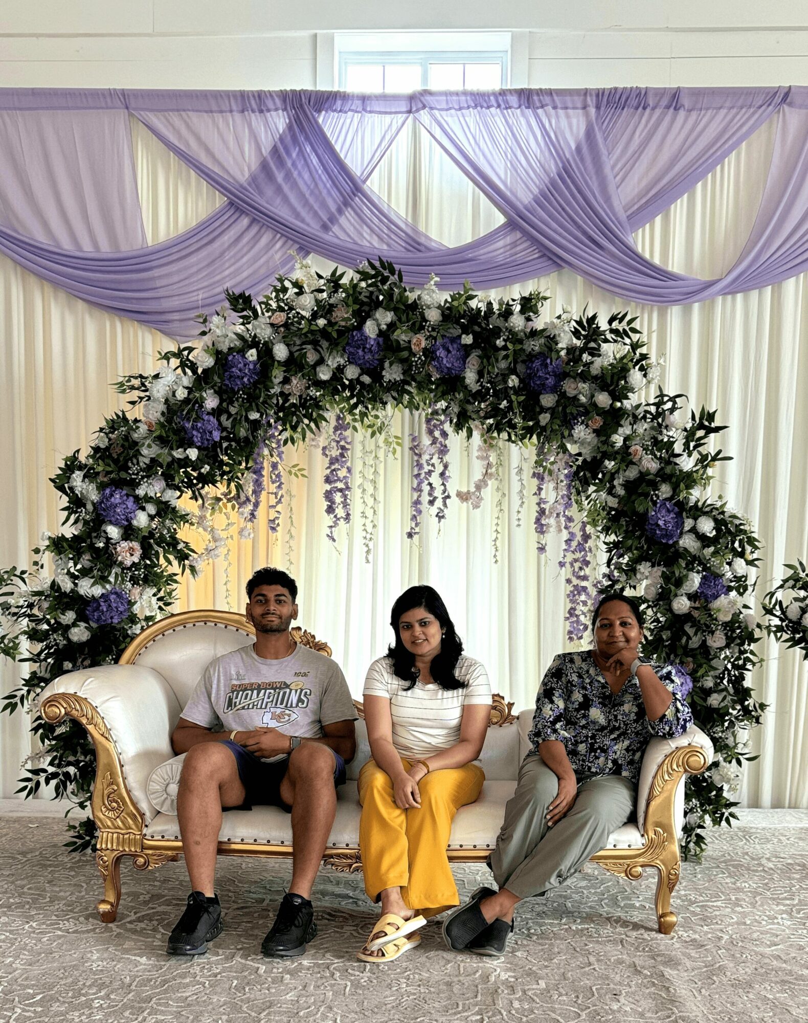 Three people sitting on a gold ornate sofa under a purple floral arch with draped purple fabric above.