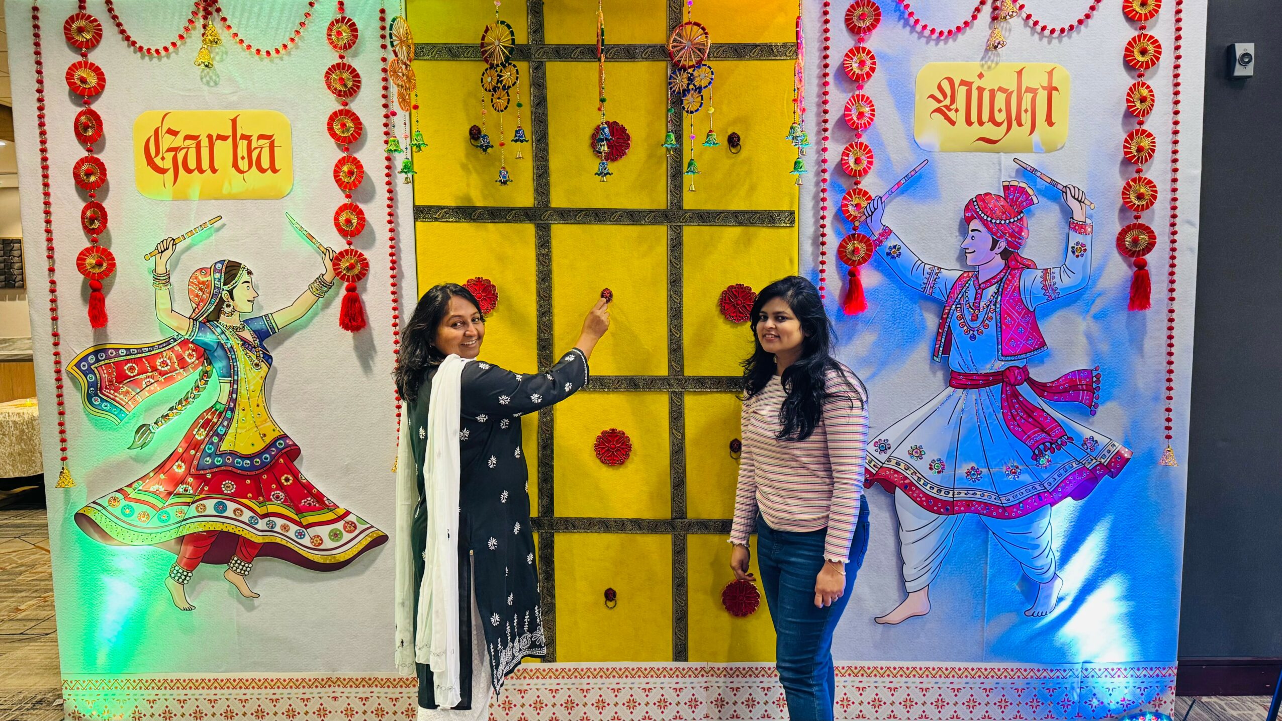 Two women stand in front of colorful decorated panels with dancers and text, smiling at the camera.