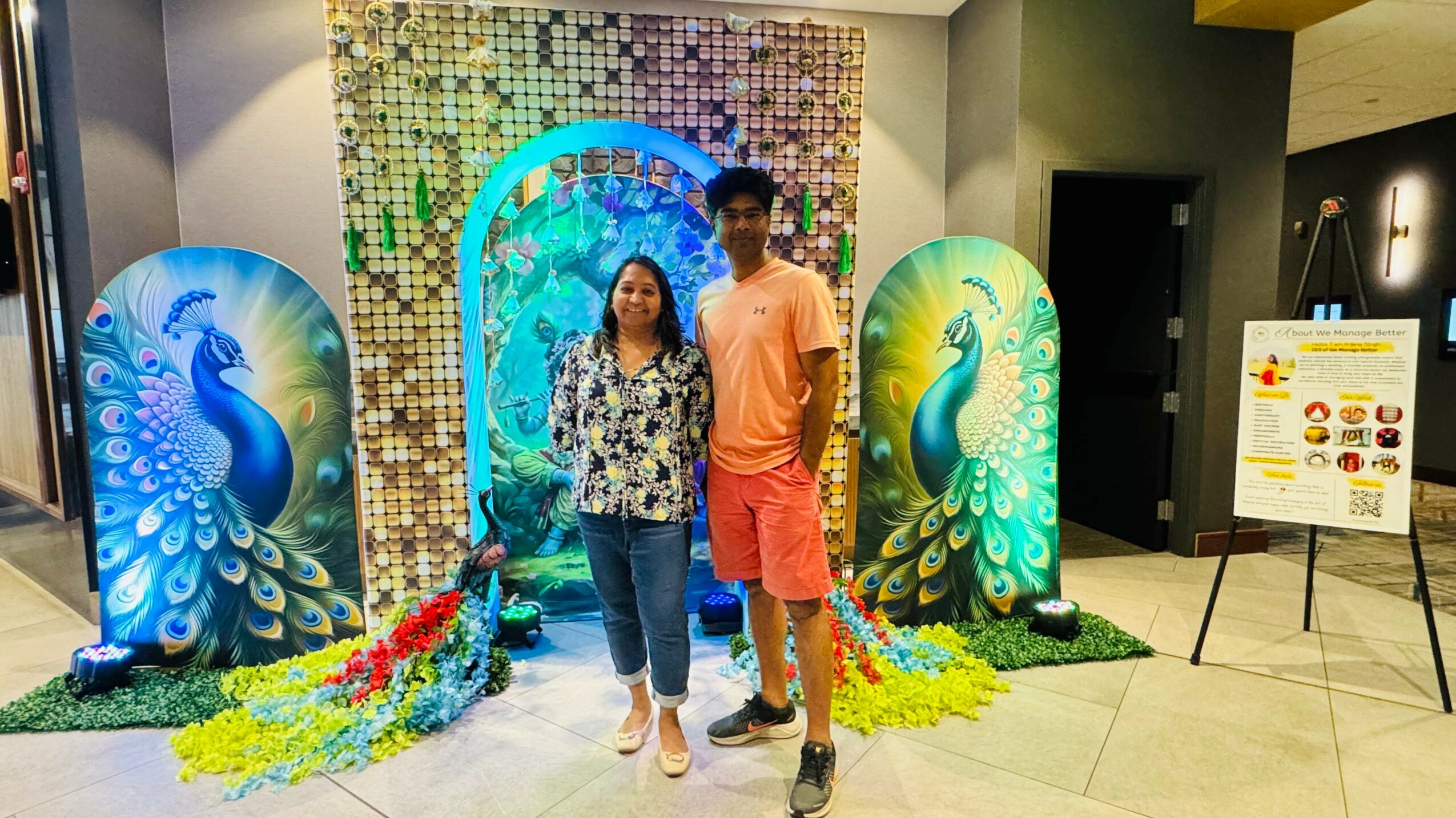Two people standing in front of peacock-themed decorations with colorful peacock images and a mosaic backdrop.