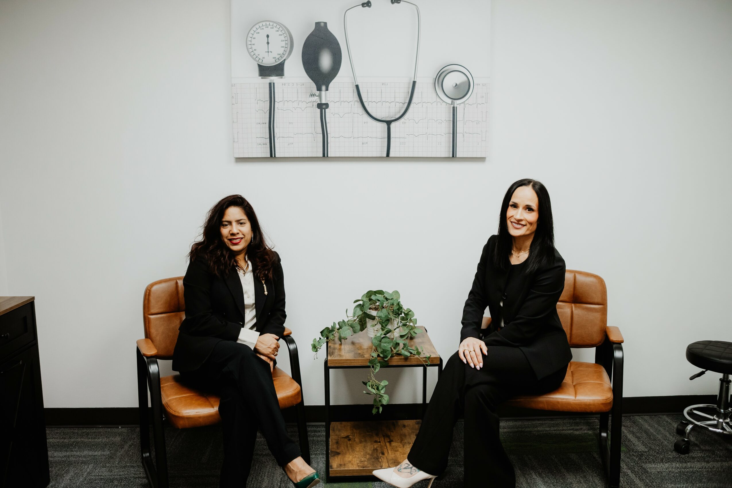 Two women sit on brown chairs in a room with a white wall and medical-themed artwork above them, a small table with a plant between them.