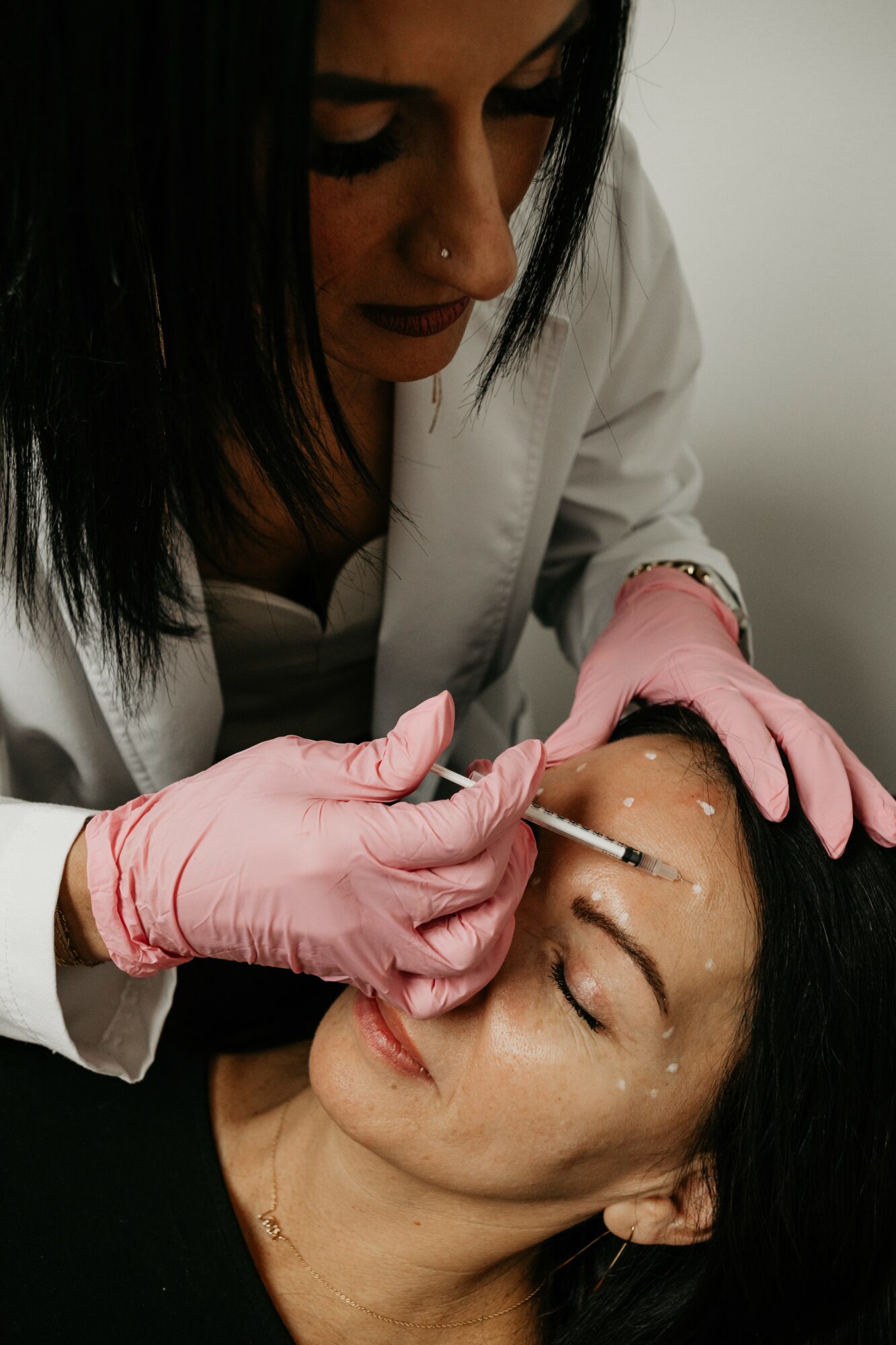 A woman in pink gloves performs a cosmetic procedure on another woman's face.