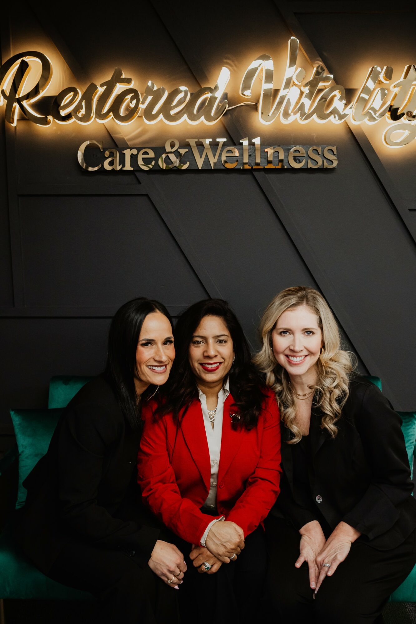 Three women sitting together in front of a sign that reads 'Restored Attitudes Care & Wellness'.